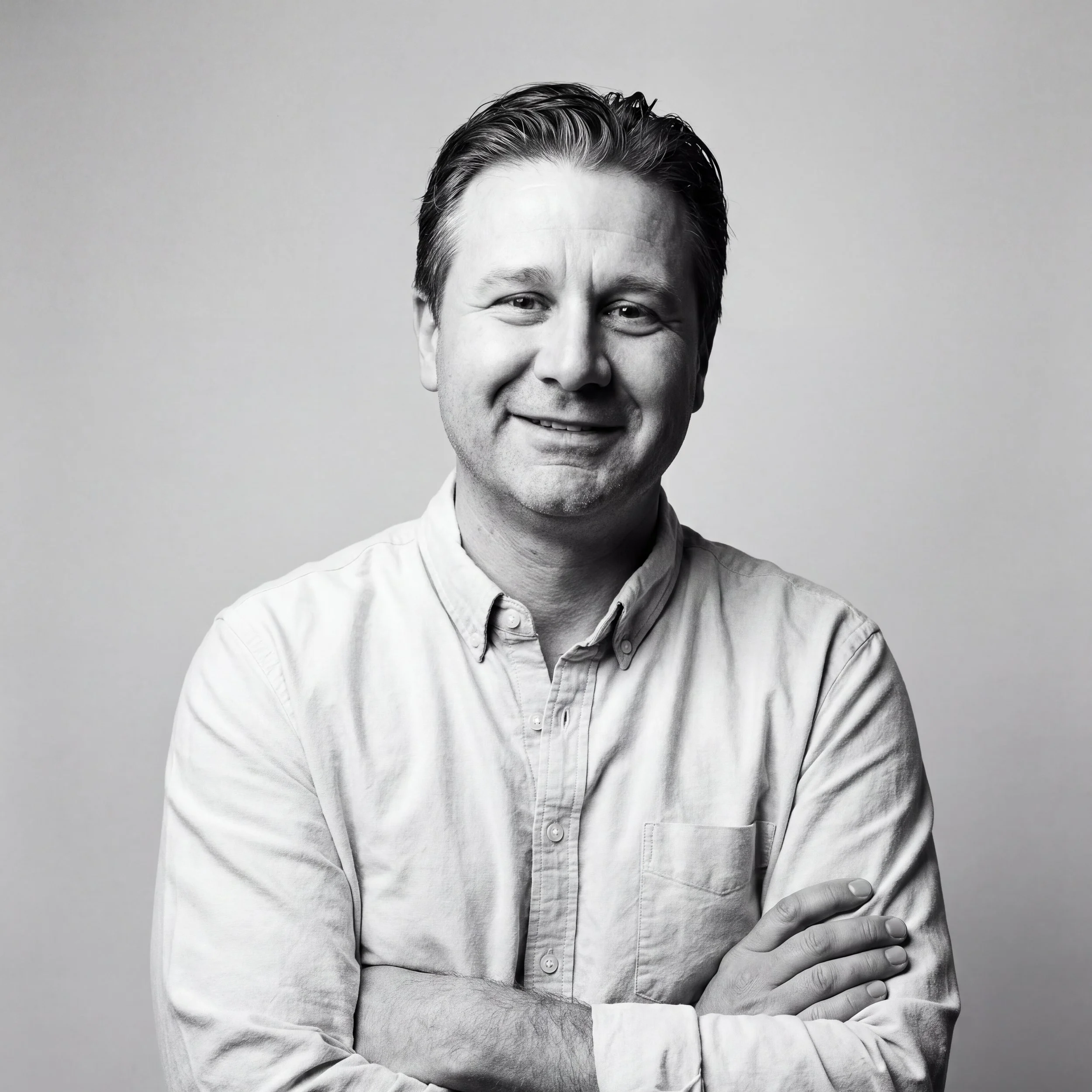 A black and white portrait of a man with dark, slicked-back hair, smiling slightly, wearing a light-colored button-up shirt with his arms crossed, standing against a plain, light-colored background.