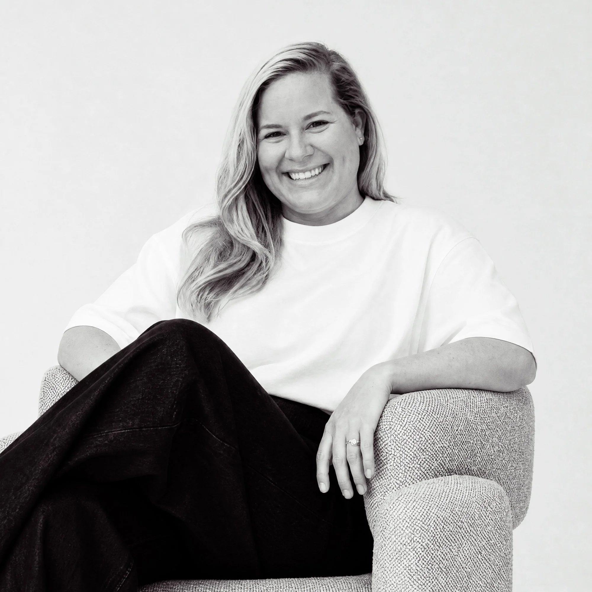 Black and white portrait of a woman smiling, sitting on a textured chair, with her hand resting on her leg, wearing a white shirt and patterned leggings.