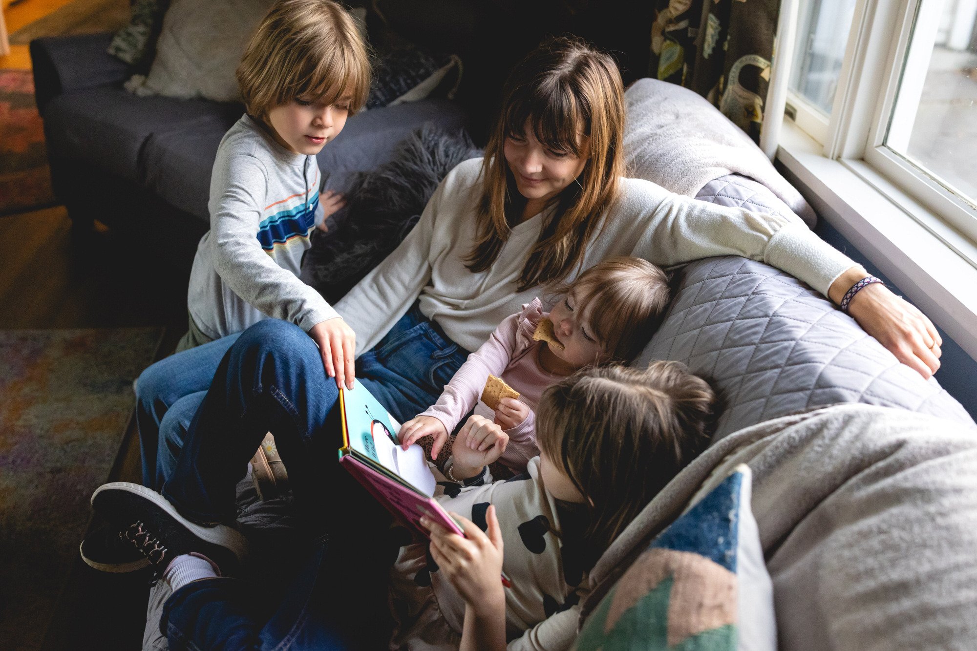 A woman and four children sitting on a gray quilted couch by a window, looking at a colorful picture book, with the children holding snacks.