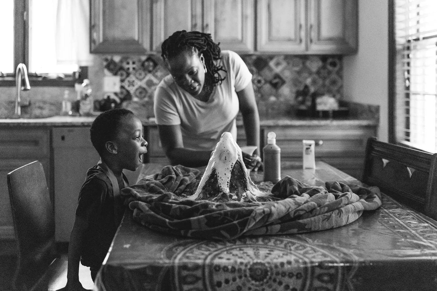 A woman and a young boy engaging in a science experiment with a bubbling volcano model on a table in a kitchen.