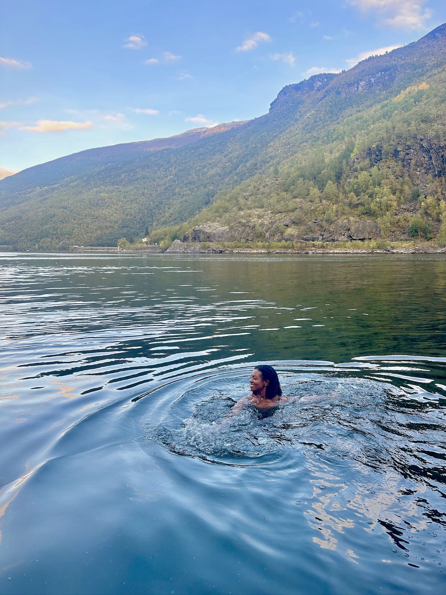 A woman swimming and smiling in a large body of water with a mountain and forested landscape in the background.