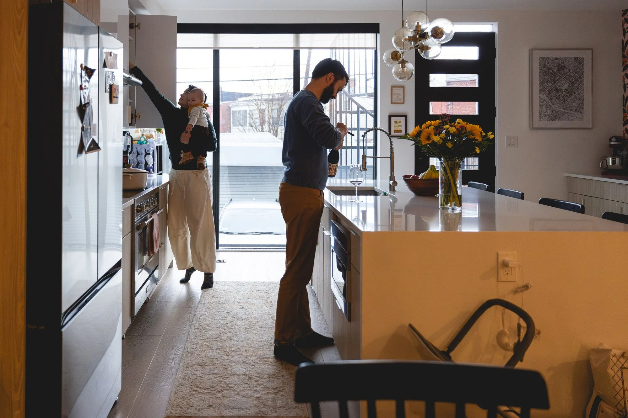 A man with a beard and glasses is standing at the kitchen island, pouring a beverage into a wine glass. Behind him, a woman is holding a baby and reaching into a cabinet. The kitchen has large windows with black frames, a floral centerpiece on the island, modern lighting fixtures, and framed artwork on the wall.