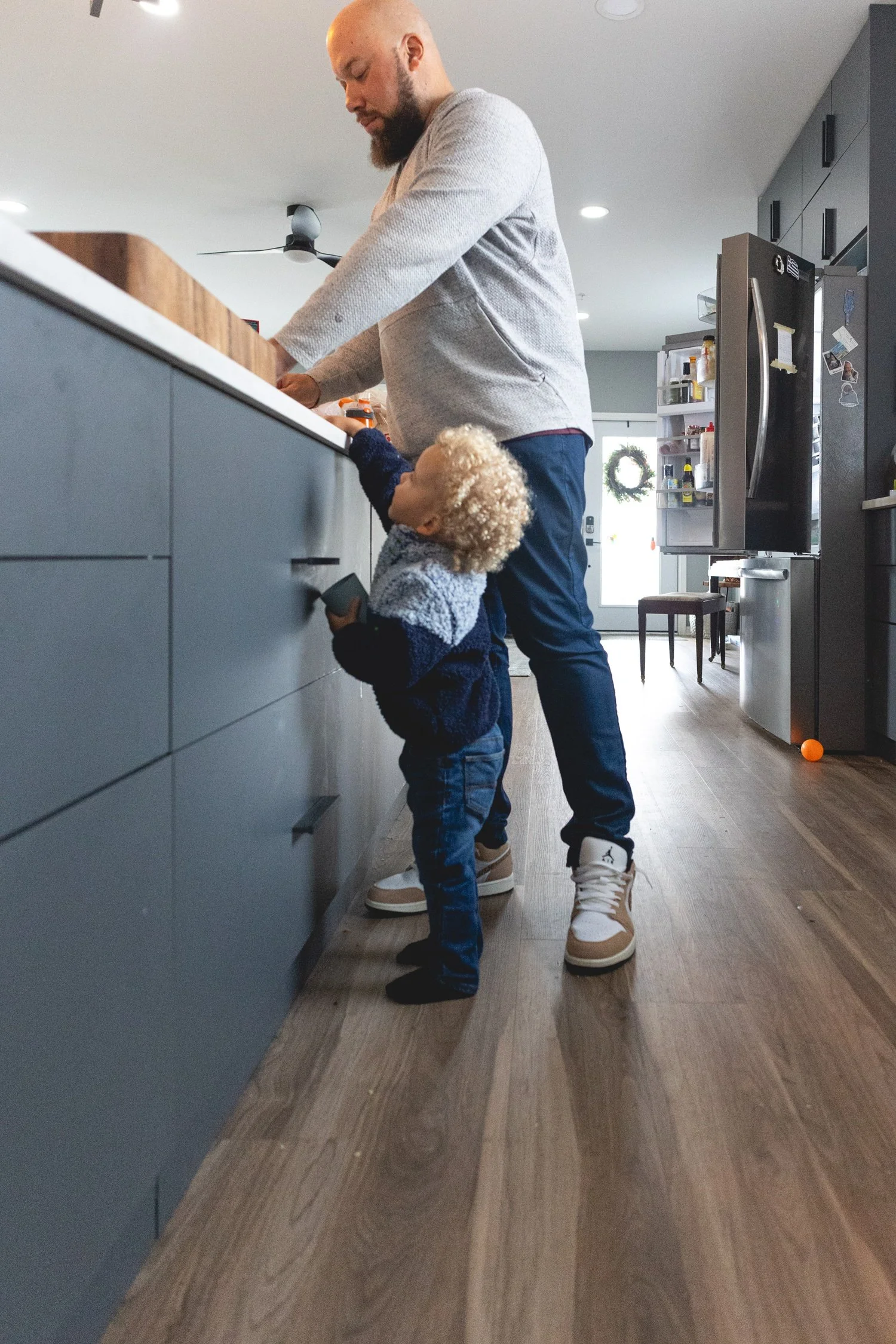 A man and a young child standing in a kitchen. The man is reaching into the refrigerator while the child looks up at him, holding an electronic device. The scene features modern kitchen cabinetry, a refrigerator, and kitchen details in the background.