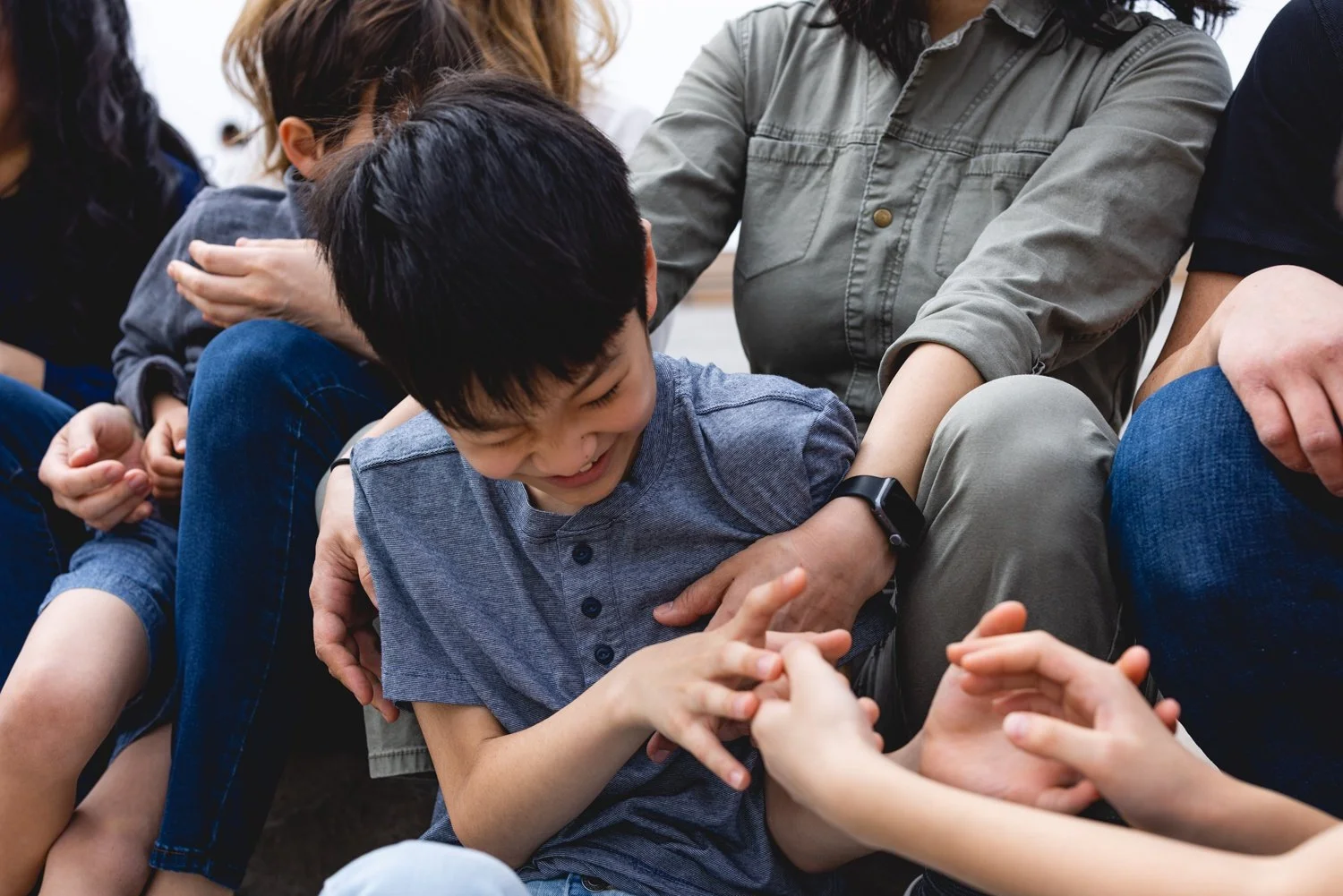Young boy laughing and playing with others in a group, sitting on the floor, touching hands with another person.