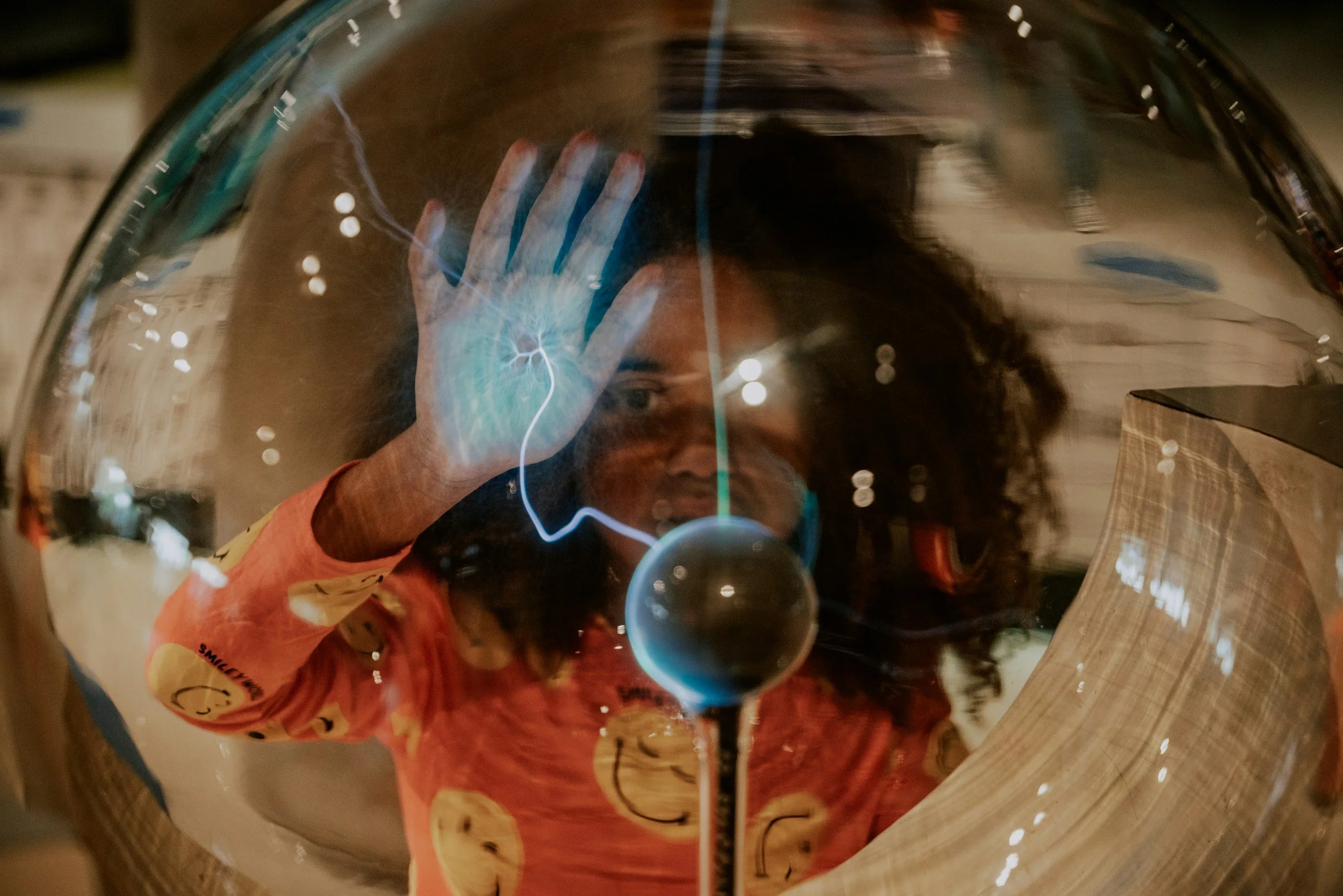 A person taking a selfie through a reflective, shiny surface, possibly a metallic or mirror-like object, with their hand raised and face partially visible.
