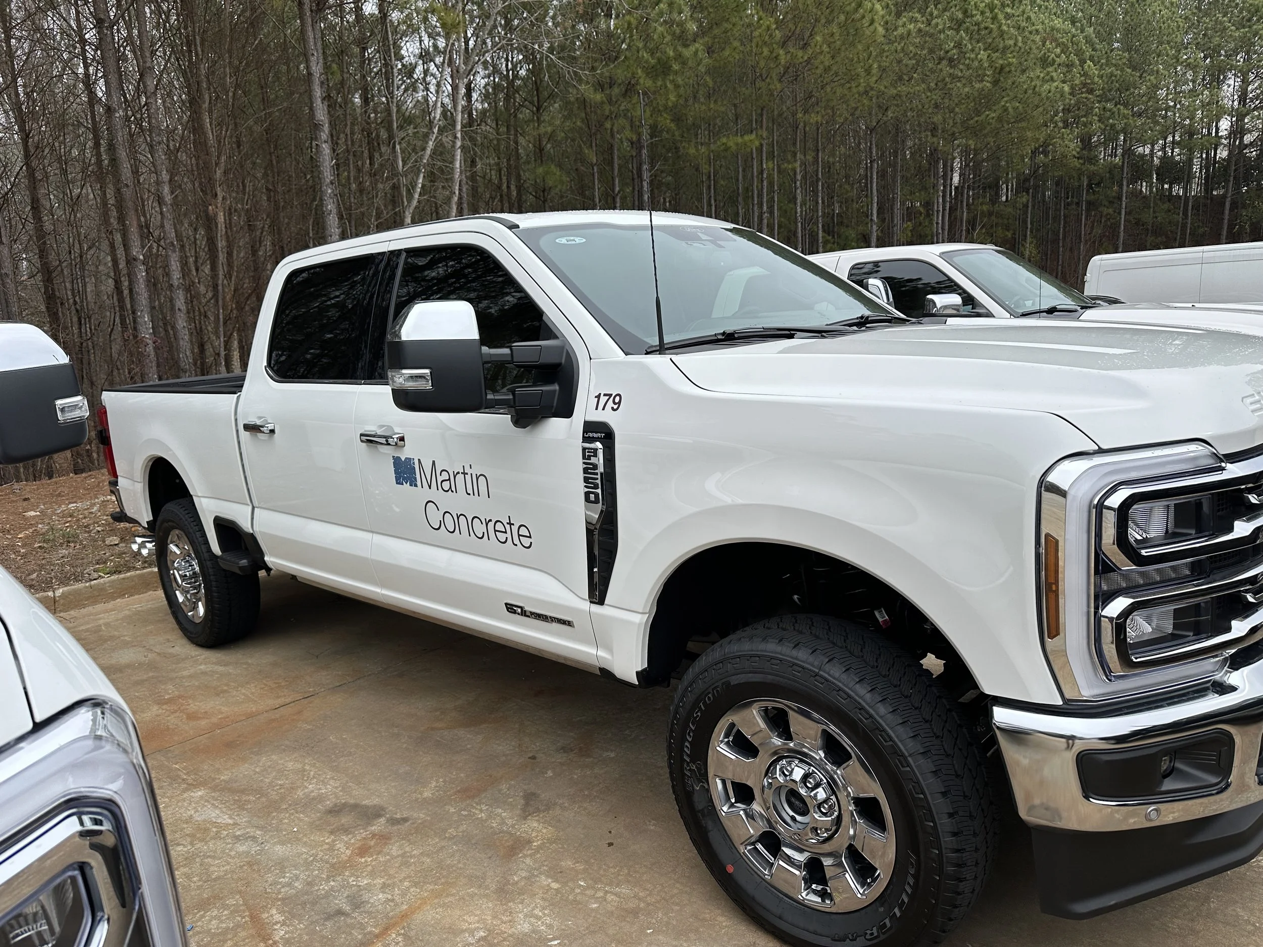 A white service pickup truck with the logo 'Martin Concrete' parked among other vehicles outdoors, with a wooded background and a cloudy sky.