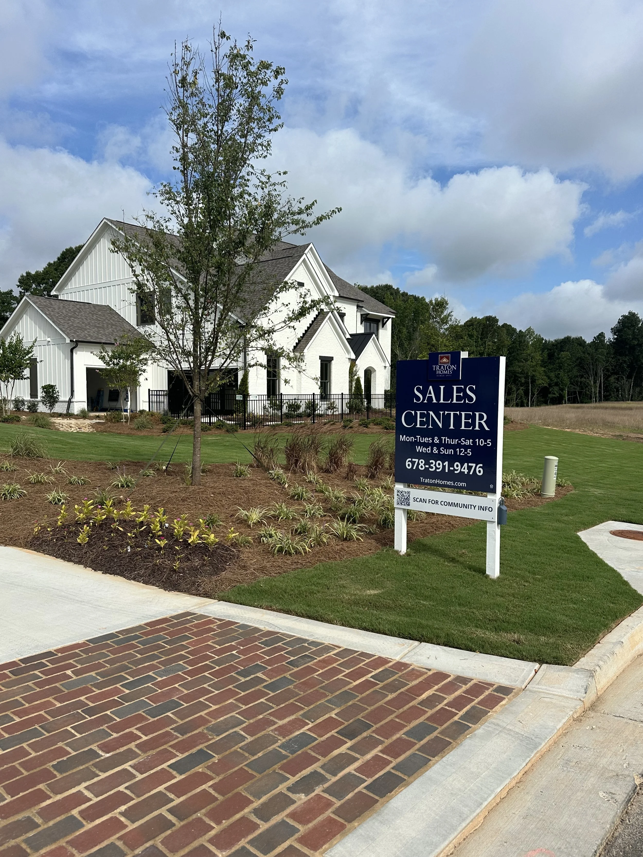 A residential real estate sales center with a white house, landscaped yard, and a sign for Traton Homes indicating their sales hours and contact information.