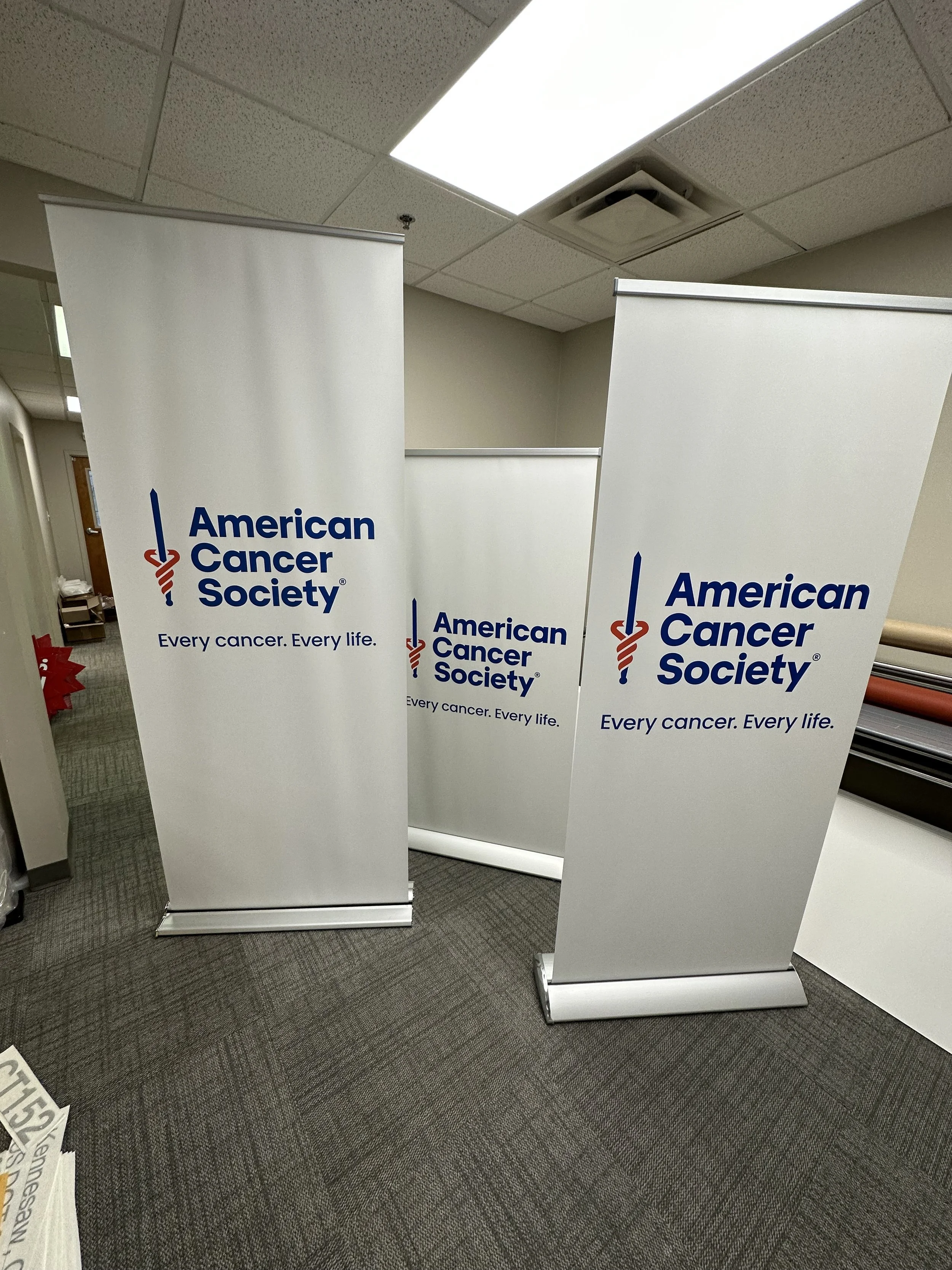 Two freestanding banners with the American Cancer Society logo and slogan placed on a gray carpeted floor in an indoor office or conference room setting.