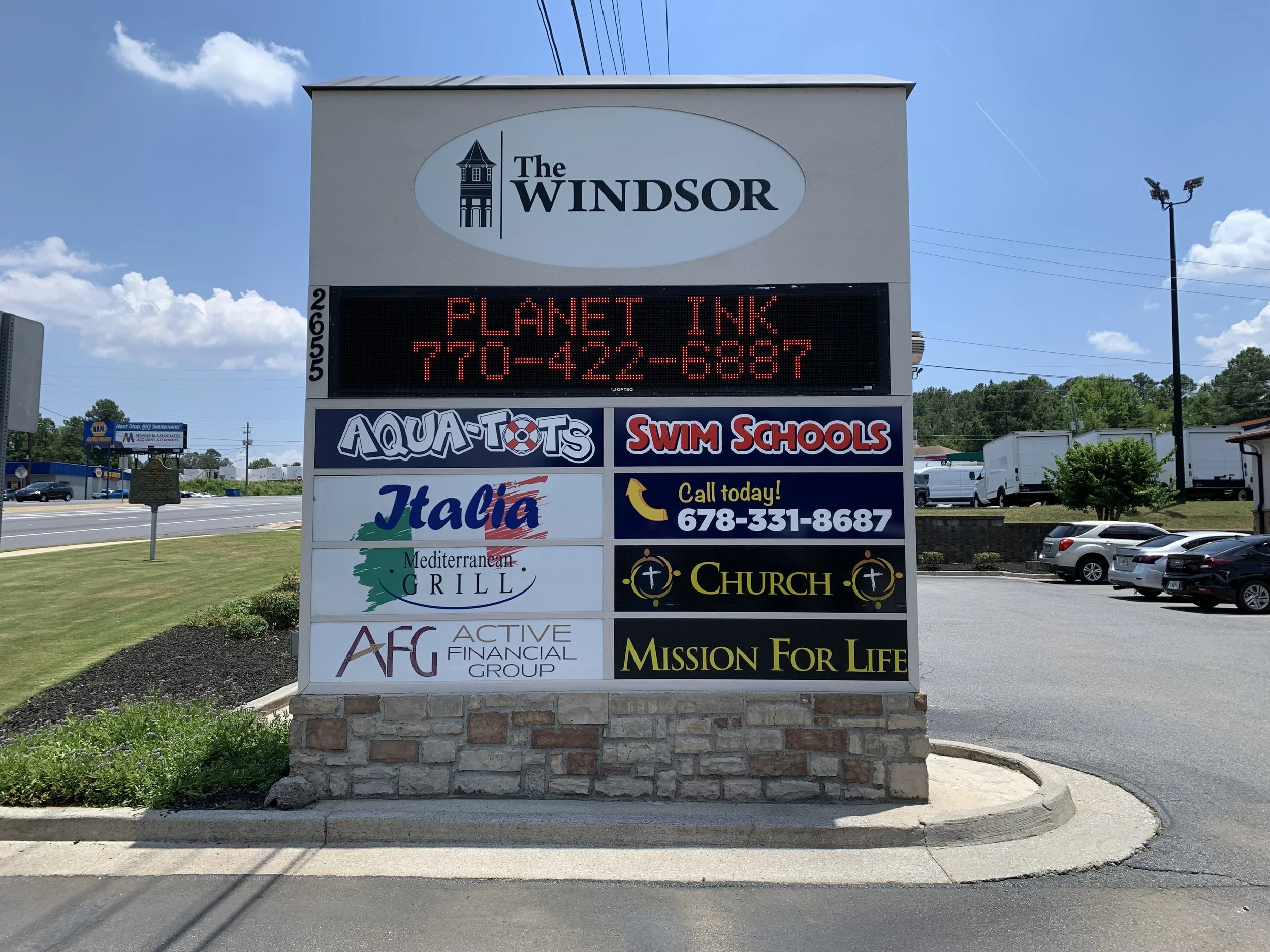 Sign for Windsor with a digital message board advertising Planet Ink and a contact number, surrounded by businesses including Aqua-Tots Swim Schools, Italic Mediterranean Grill, and others, set in a parking lot under a bright blue sky with scattered clouds.