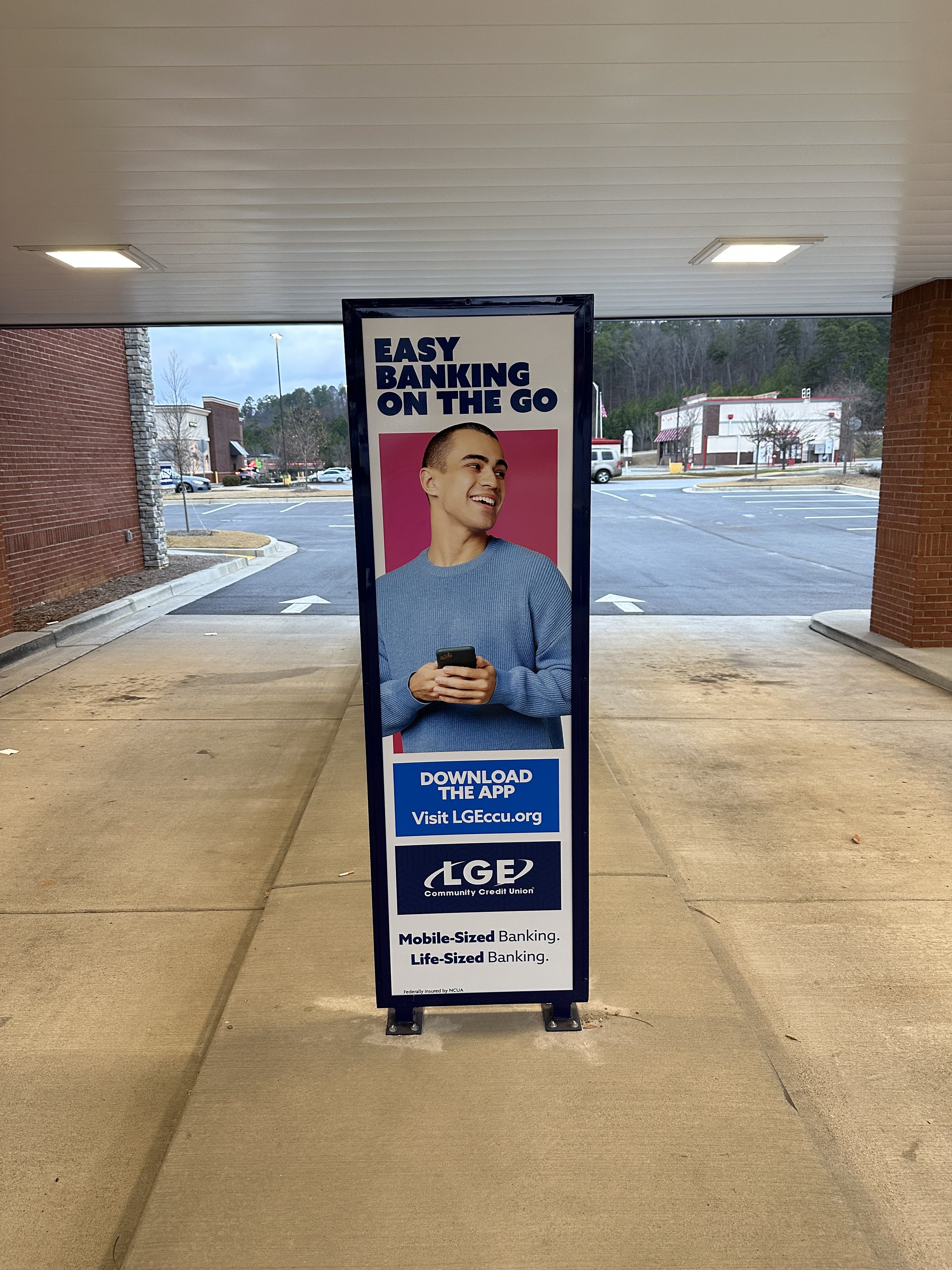 A freestanding sign outside of a community credit union promoting mobile-sized and life-sized banking options with a picture of a smiling person holding a phone. The sign includes the logo of LGE Community Credit Union and a website link.