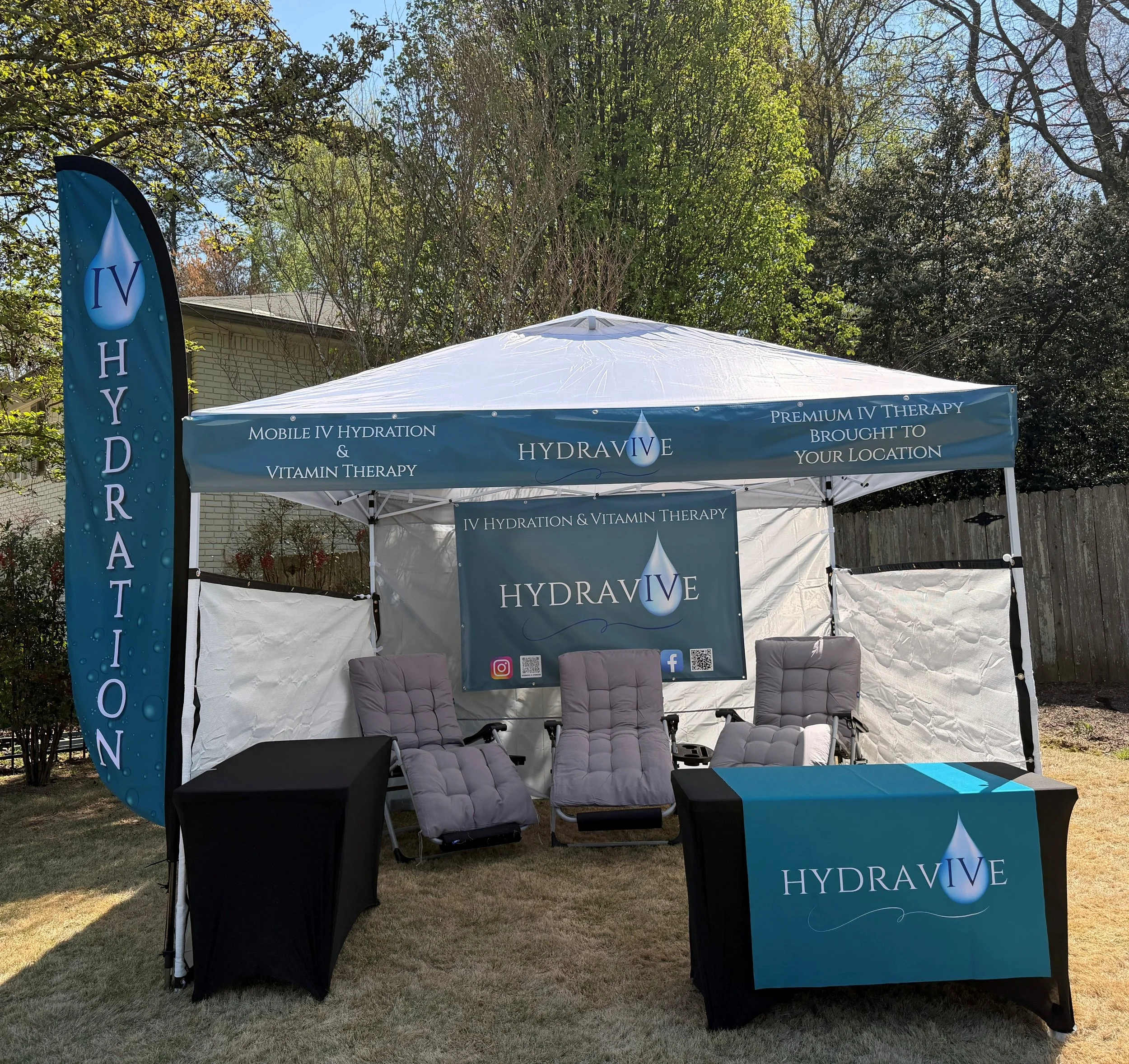 Outdoor event booth with blue and white Hydravive banner, promoting IV hydration and vitamin therapy, set up under a white canopy tent with chairs in front, surrounded by trees and a house in the background.