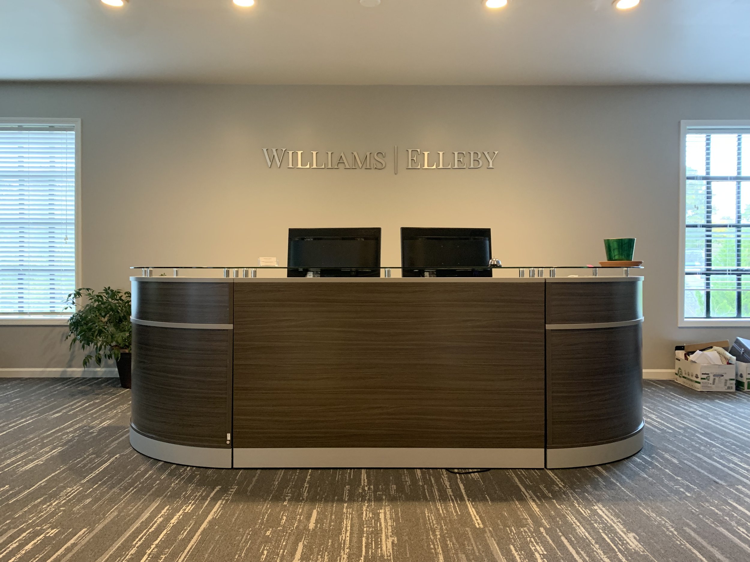 Reception desk with two computer monitors, a green bowl on a tray, plants on the side, and the words 'WILLIAMS | ELLBEY' on the wall in the background, with windows on either side of the wall.