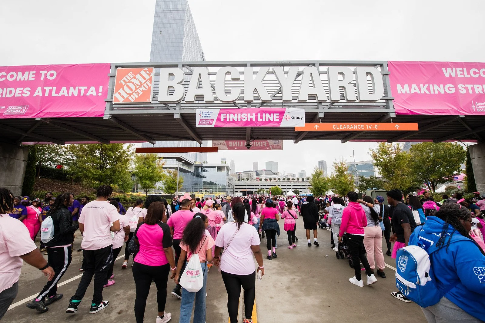 Crowd of people in pink, purple, and black clothing walking under a large metal archway with a pink banner that says 'Welcome to Making Strides Atlanta!', in front of a cityscape with tall buildings. The archway has a sign for a finish line and a clearance height of 14 feet.