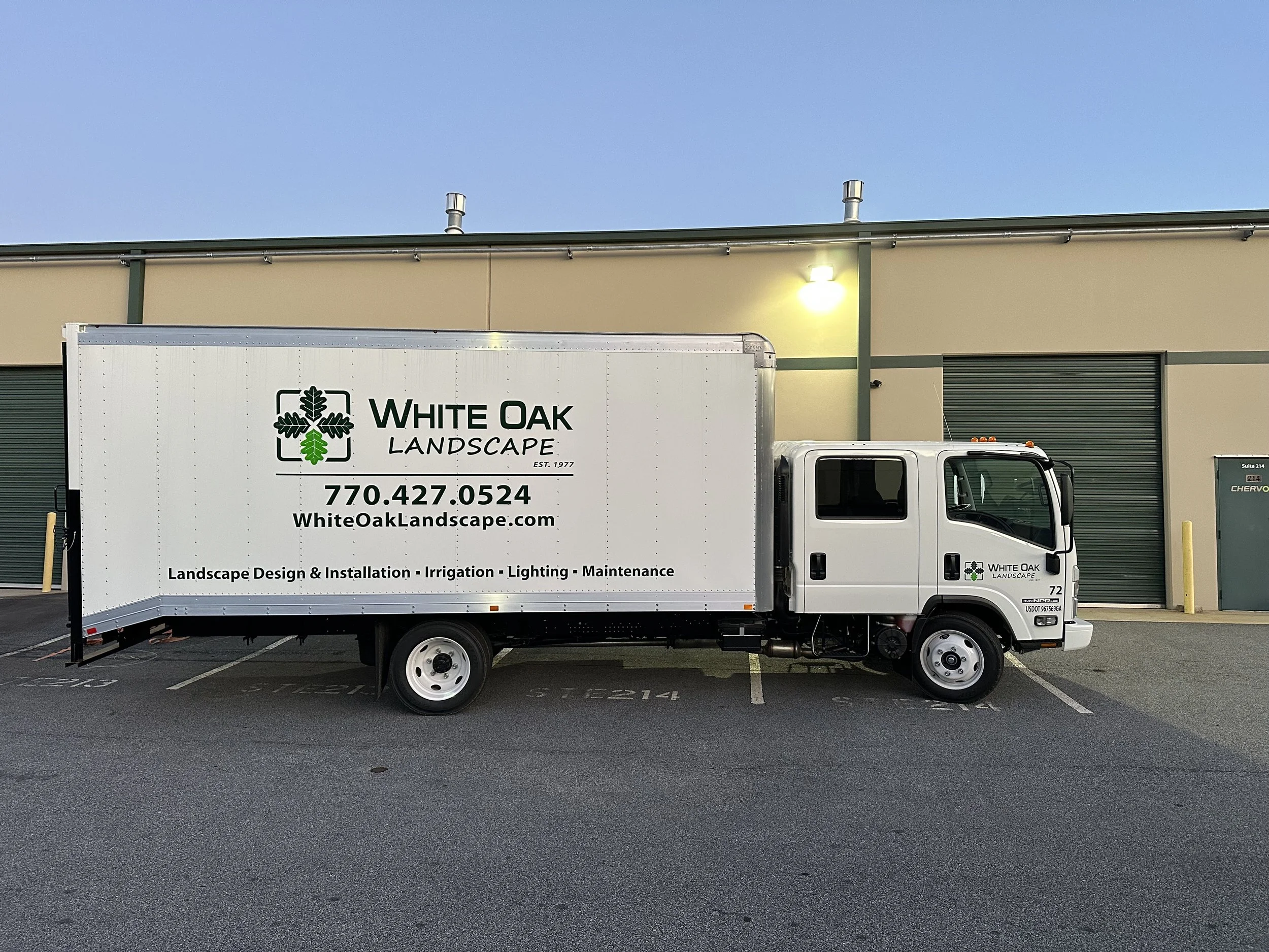 A White Oak Landscape delivery truck parked in a parking lot in front of a beige industrial building with green trim and closed rolling doors. The truck has a logo with a tree and some text, and is positioned to the right of the image. The sky is cle