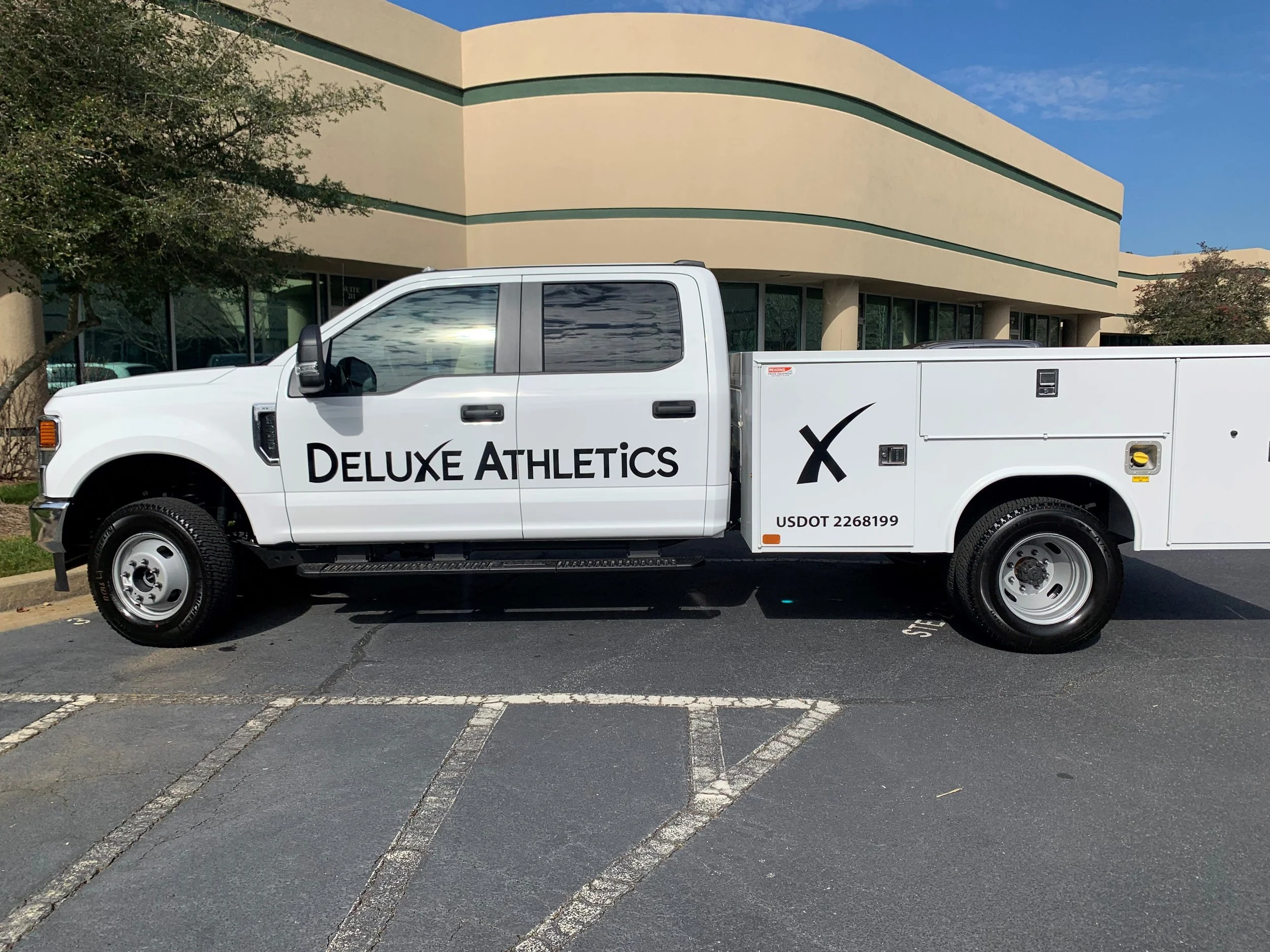 A white pickup truck with 'Deluxe Athletics' written on the side is parked in a parking lot in front of a beige building with green accents. The sky is partly cloudy, and there are some trees on the left and right sides of the building.