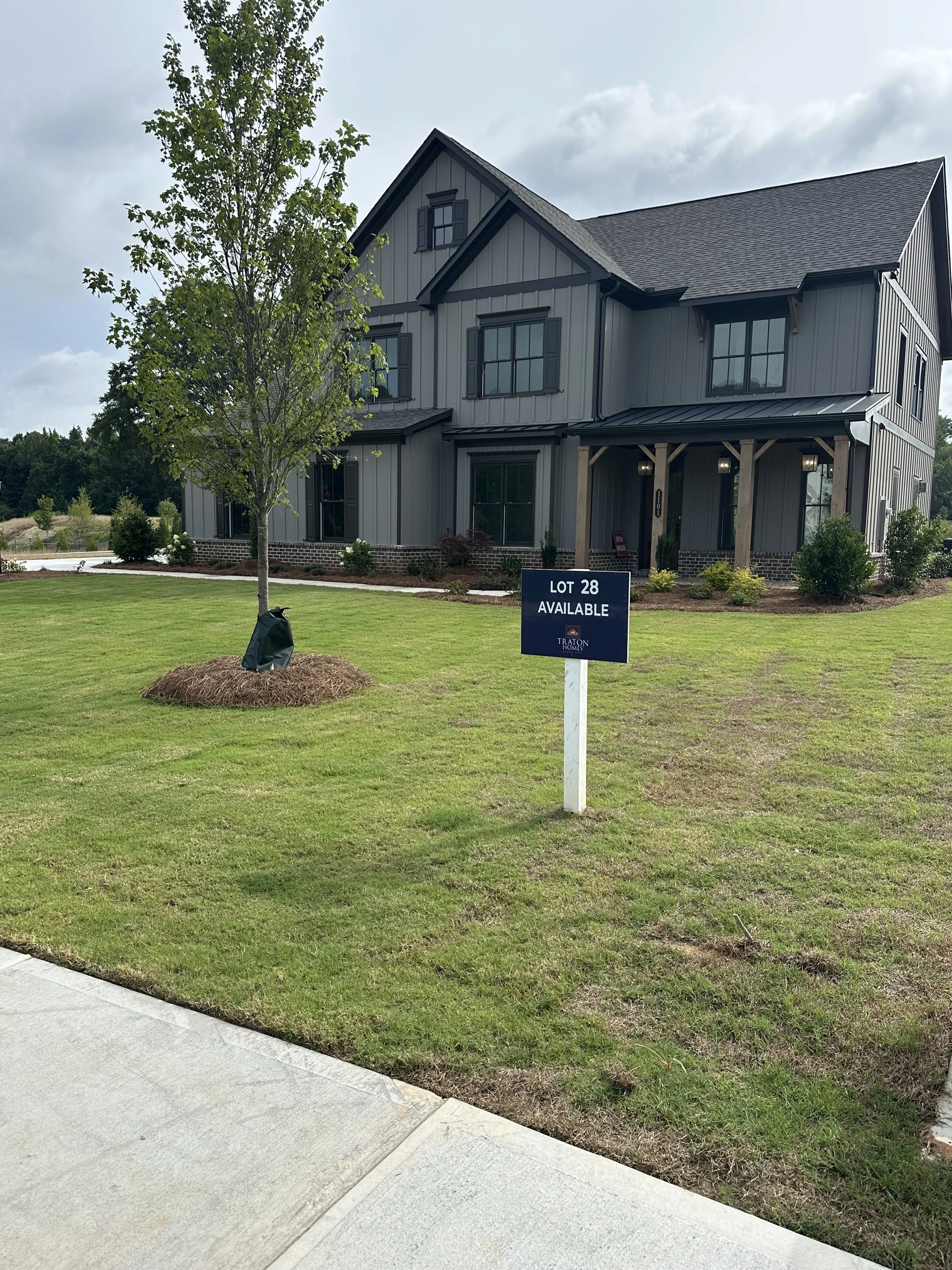 New two-story gray house with black shutters and a small front porch, surrounded by landscaped greenery, under a cloudy sky, with a sign indicating lot 28 available for sale.