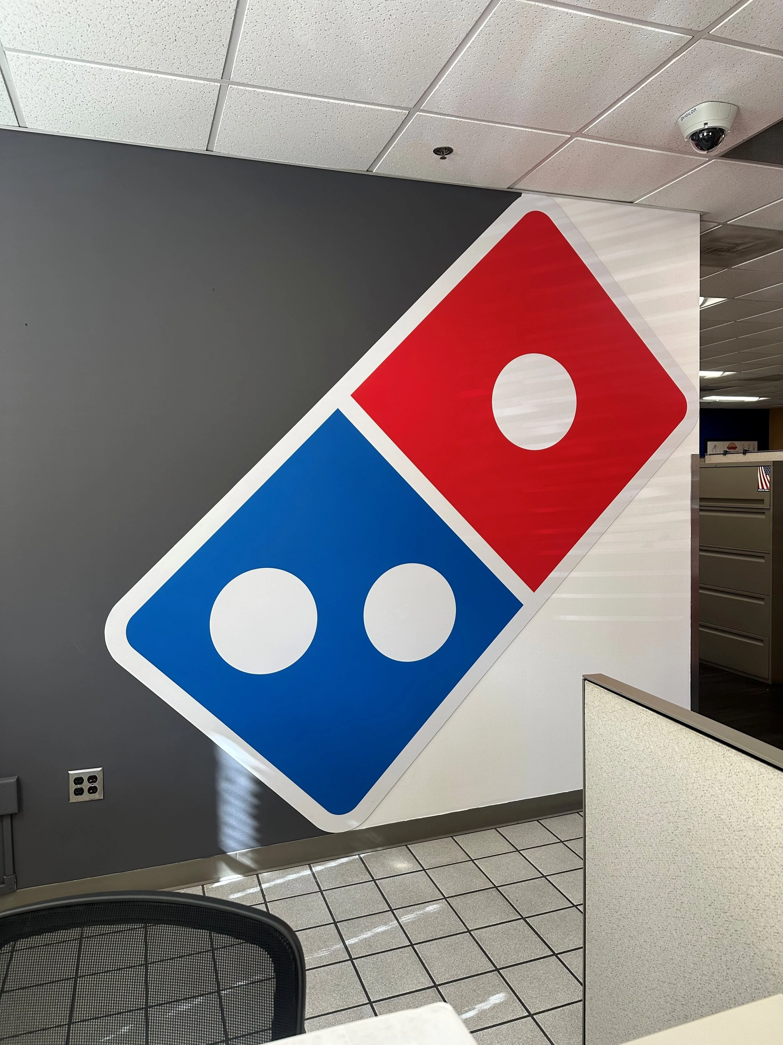Close-up view of a Domino's Pizza logo sticker on a wall at an office or commercial space. The logo features a red and blue domino tile with white circles.