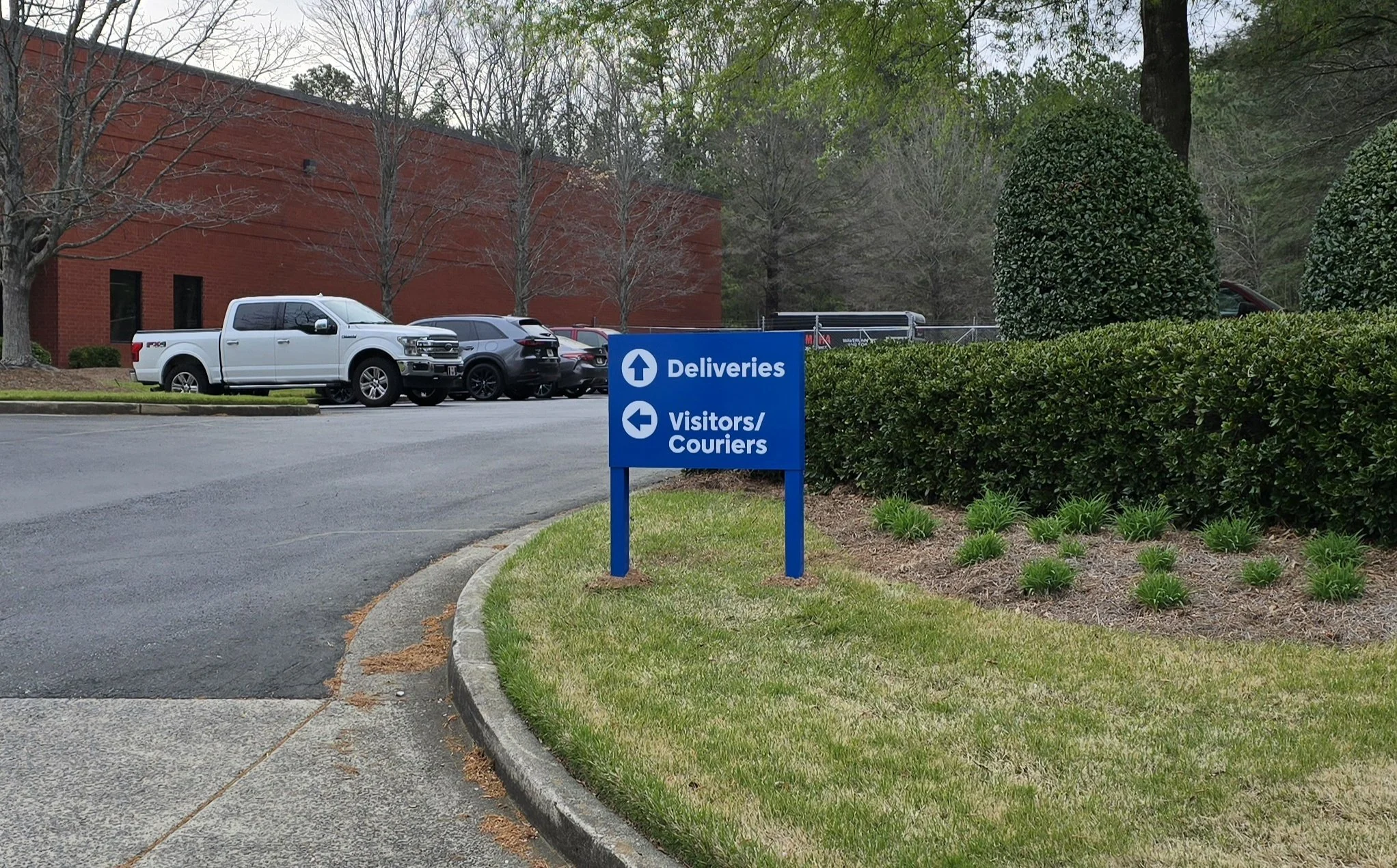 Parking lot with several vehicles parked near a red brick building, surrounded by trees with some leaves missing, and a blue directional sign indicating 'Deliveries' straight ahead and 'Visitors/Couriers' to the left.