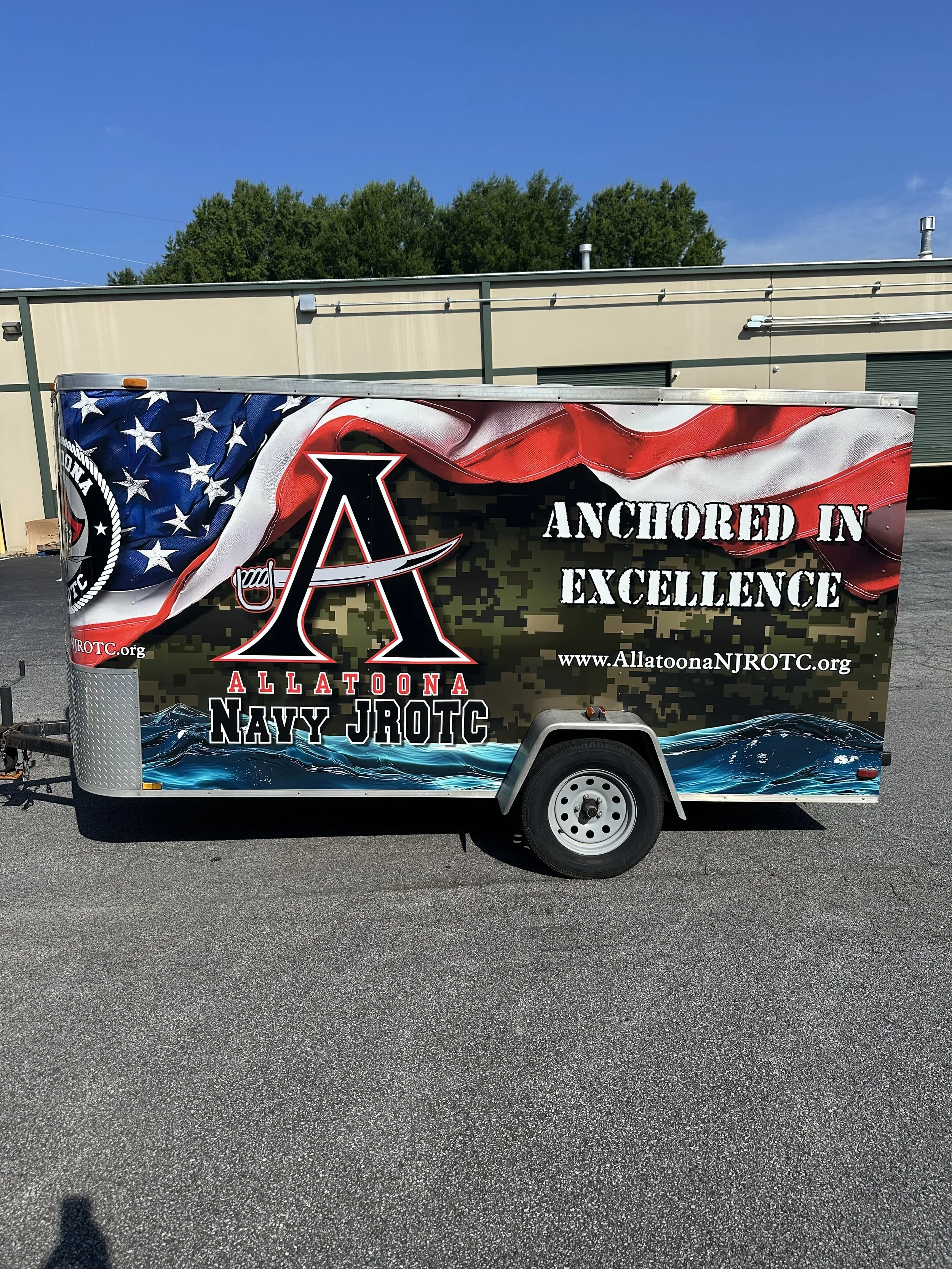 A trailer decorated with the American flag and military-themed graphics, displaying the text "Anchored in Excellence" and the website www.AllatoonaNJROTC.org.