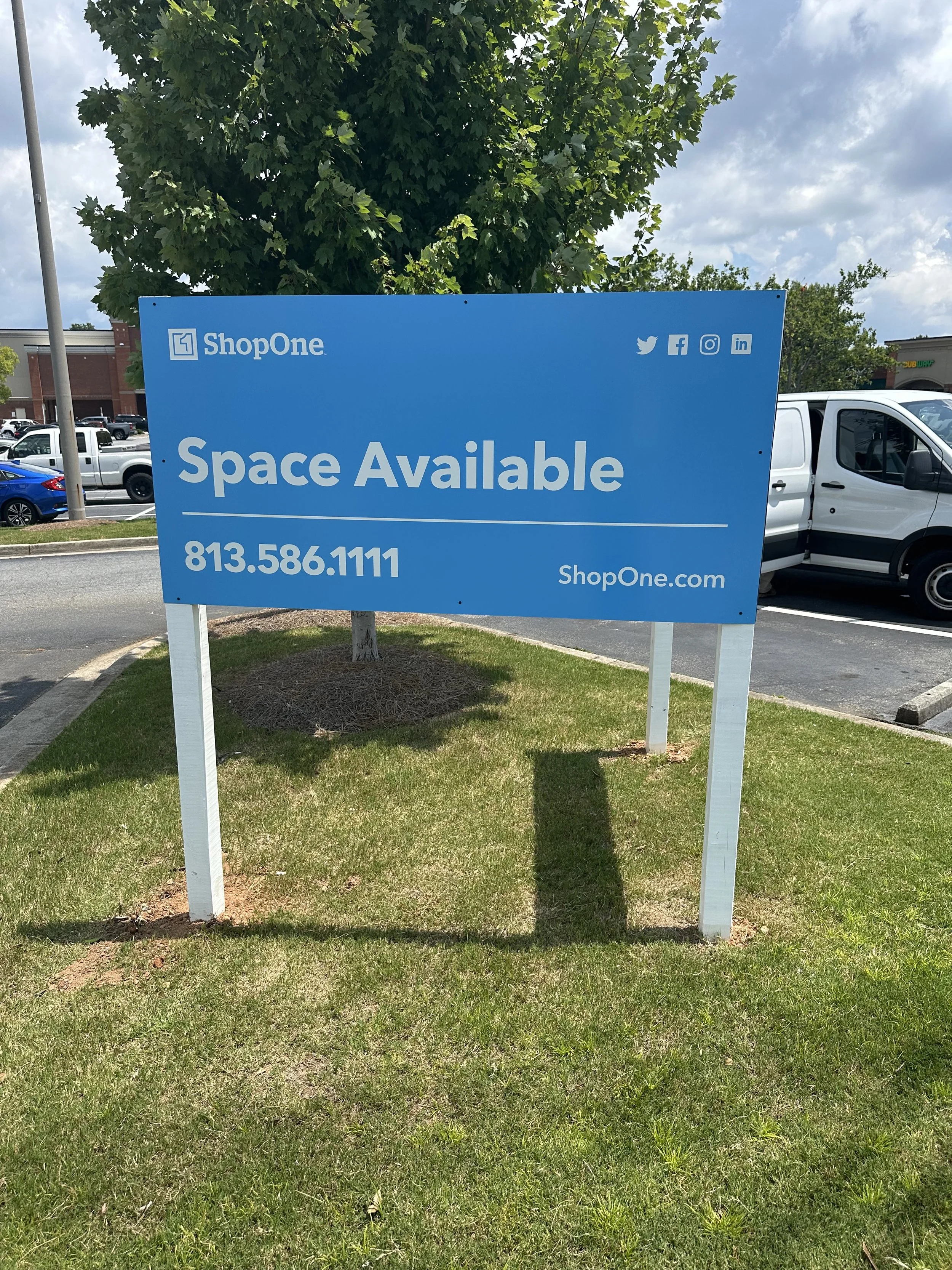 A large blue sign in a parking lot reads "Space Available" with a phone number, and the store name "ShopOne" with social media icons. There are cars and a building in the background under a partly cloudy sky.