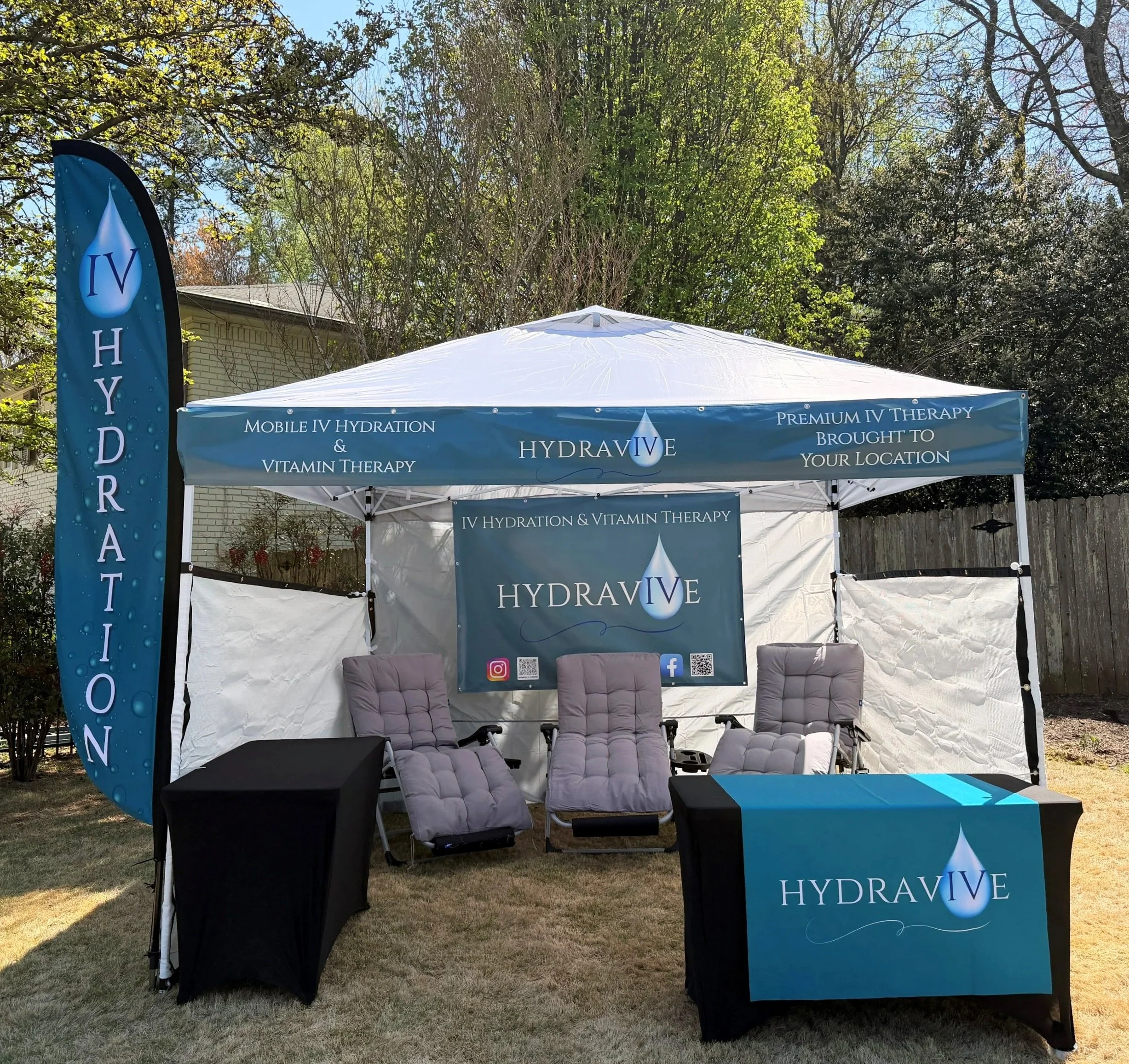 An outdoor promotional tent for Hydravive IV therapy and vitamin therapy, with chairs underneath and banners displaying the company's logo and services, set in a backyard with trees.