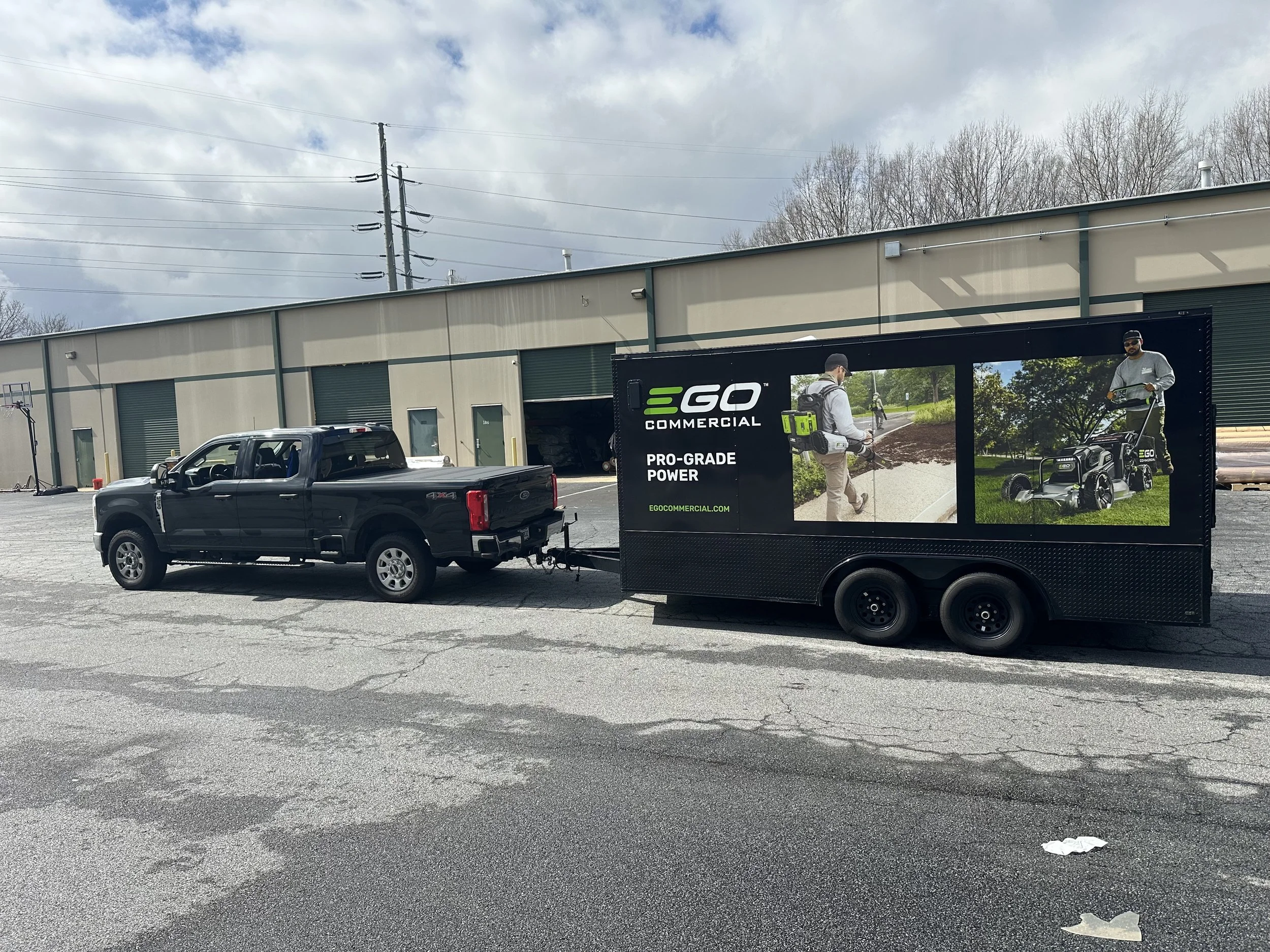 A parking lot with a black pickup truck and a mobile billboard trailer advertising EGEO Commercial, showing two men with lawn equipment, one using a string trimmer and the other using a lawnmower, with a beige industrial building and power lines in t