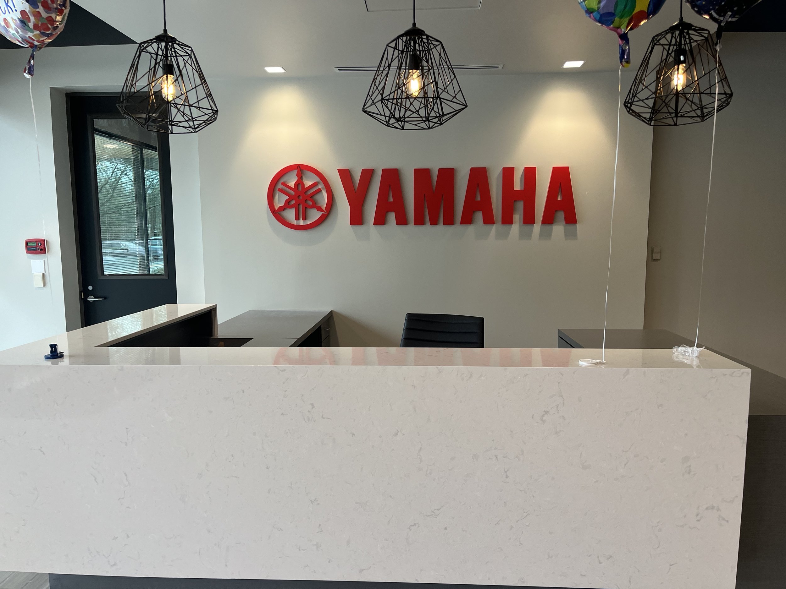 Interior of a Yamaha office reception area with a white counter, black chairs, a window, and a wall featuring a large red Yamaha logo and brand name. Overhead modern pendant lights and some colorful balloons are visible.