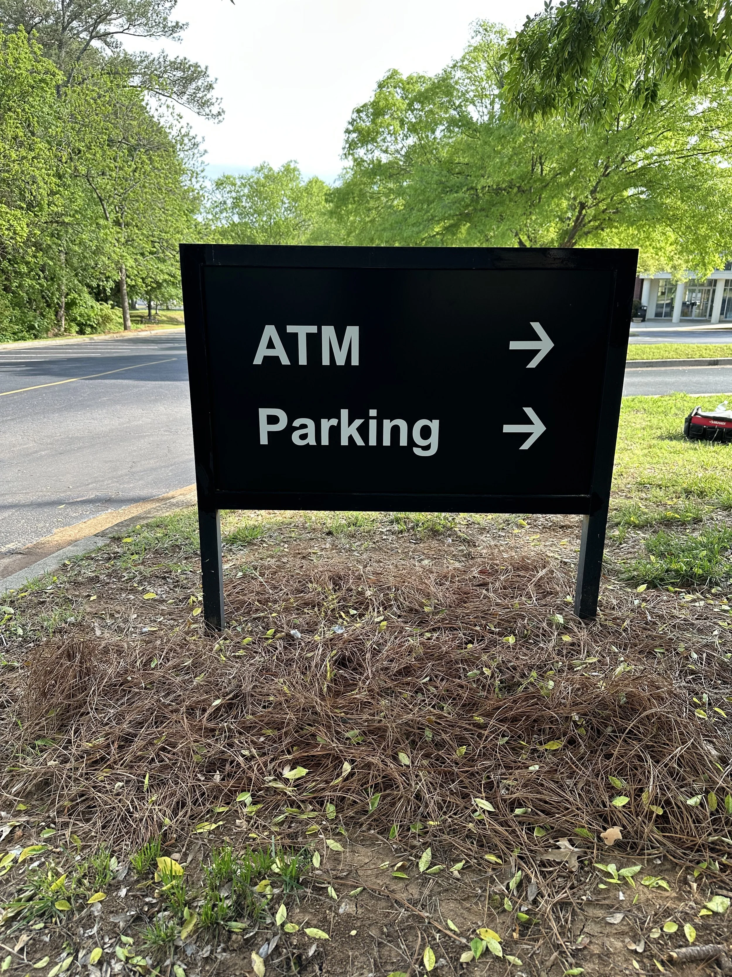 A black sign with white text and arrows pointing right, indicating the direction to the ATM and parking area, placed on a patch of ground with dry grass and some small green plants.