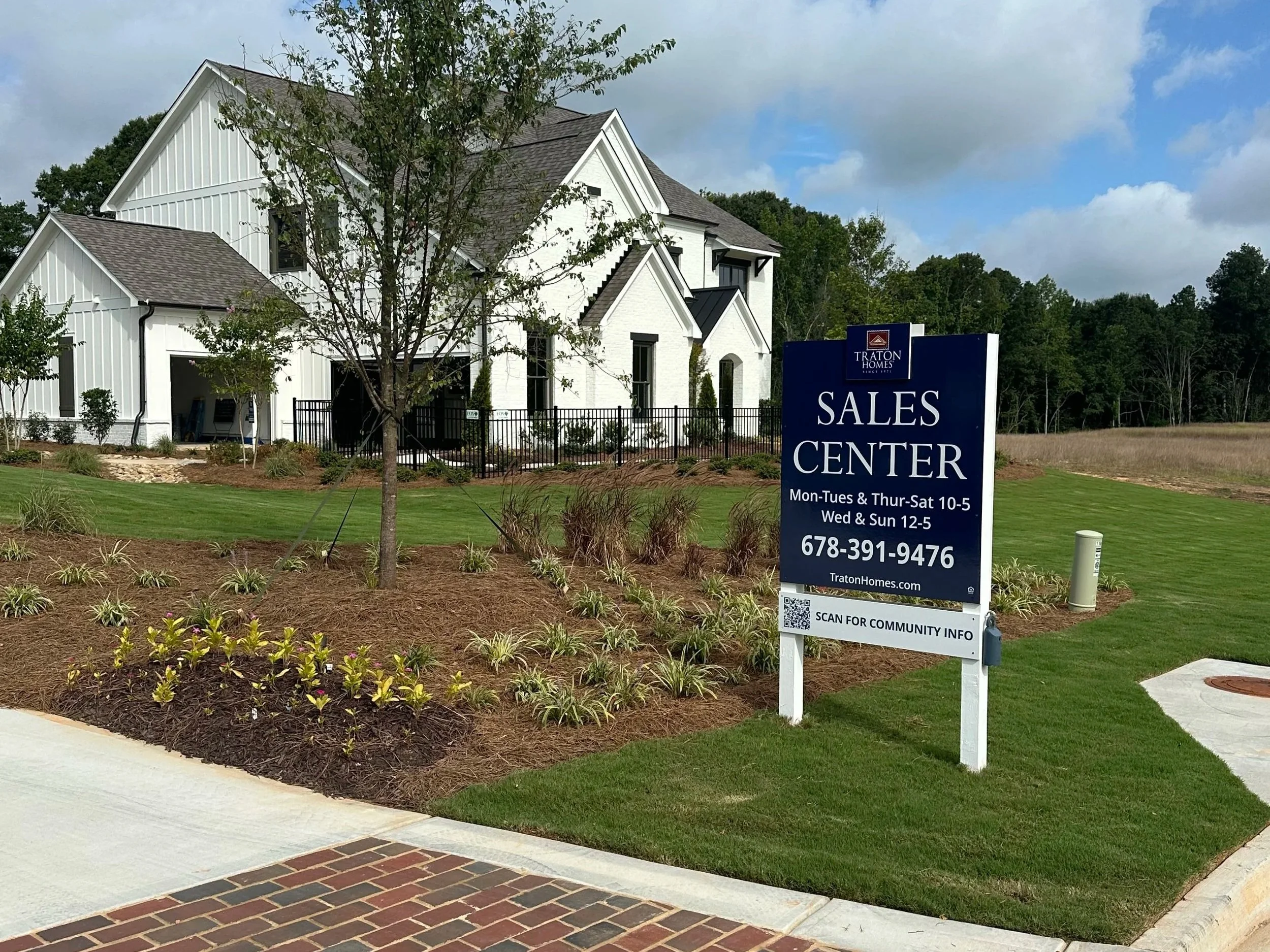 A white house with a gray roof and black window frames behind a sign that reads 'Sales Center' for Traton Homes. The sign includes office hours and a contact number. There is a landscaped area with small plants and a walking path in front of the house, under partly cloudy skies.