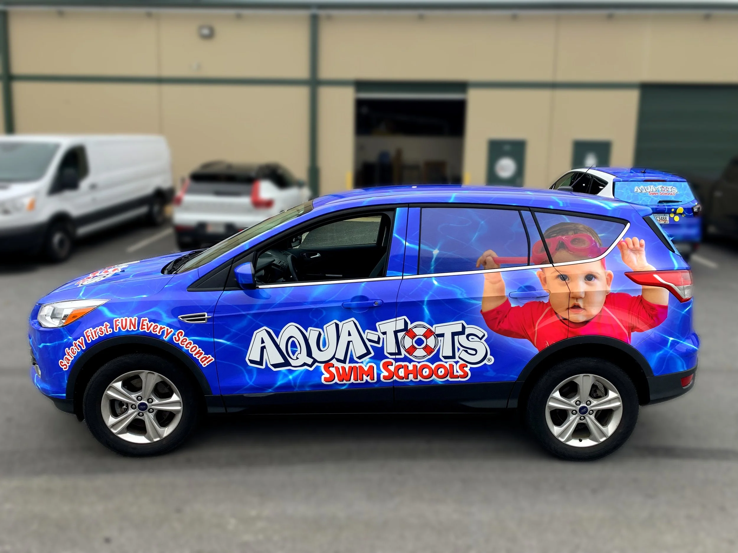 A blue Audi car decorated with graphics promoting Aqua-Tots Swim Schools, featuring a smiling child in goggles and swimwear, parked outdoors with other vehicles and a beige building in the background.