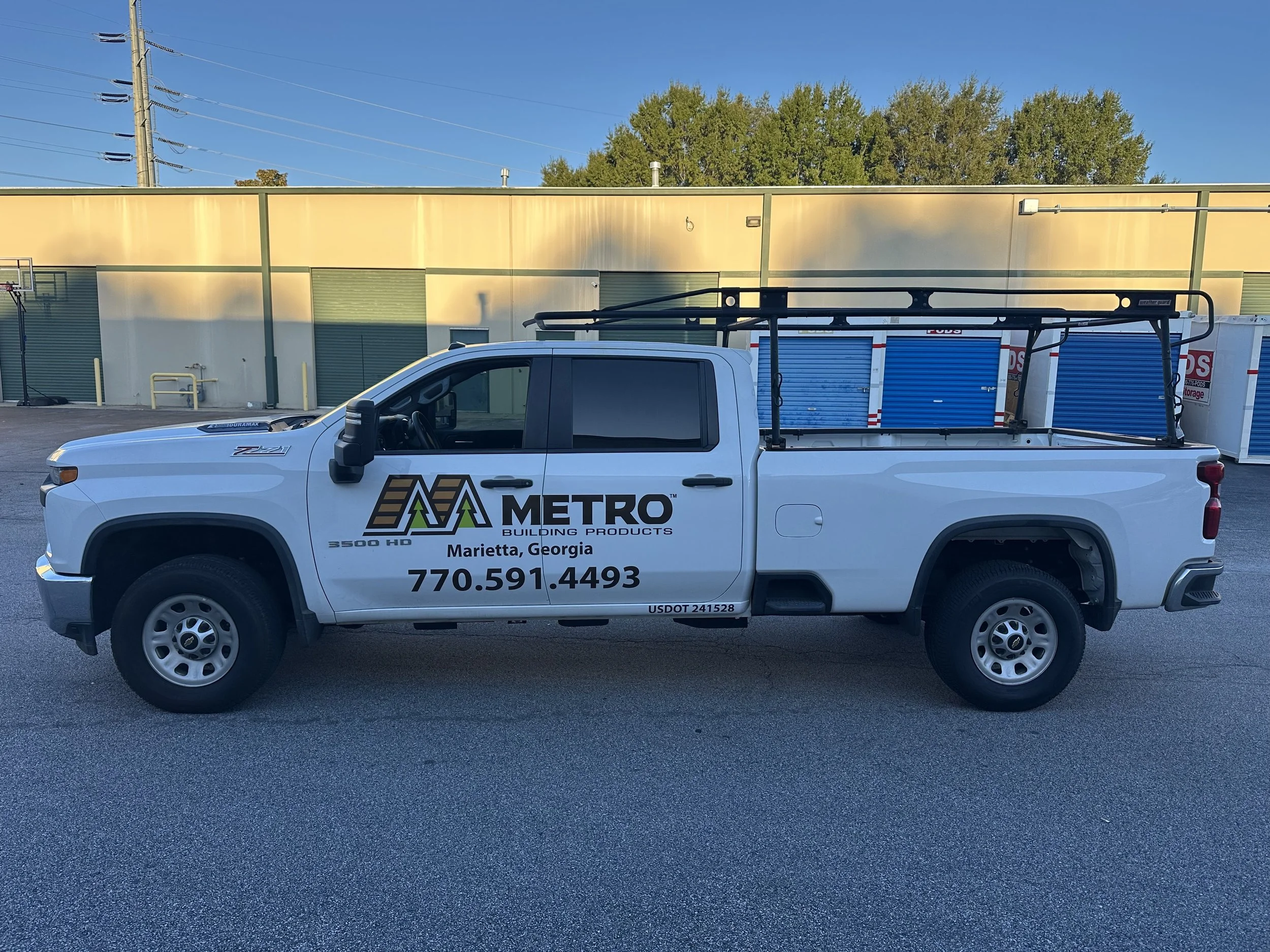 White pickup truck parked in front of a yellow building, with a large ladder on the truck's bed, lettering on the side of the truck indicating it is from Metro Building Products in Marietta, Georgia, and a soccer goal and storage units in the backgro
