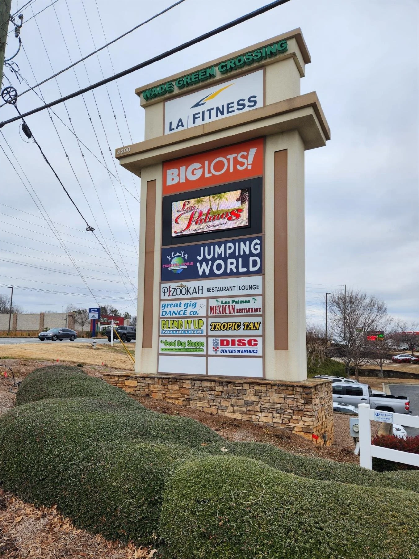 A tall signpost with multiple business advertisements along a sidewalk, including fitness, restaurants, dance, nutrition, pet store, disc golf, and community centers, under a cloudy sky.