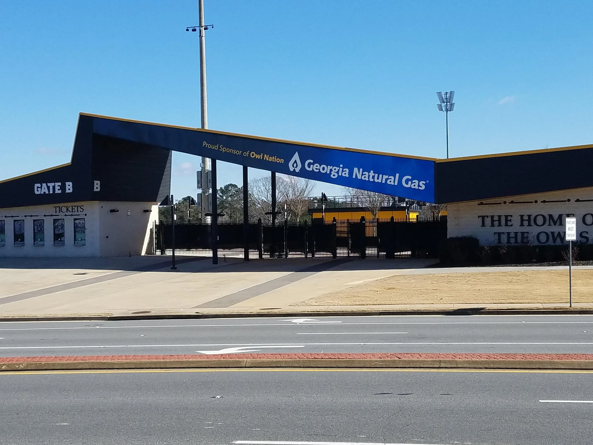 View of a stadium entrance with signs for Georgia Natural Gas, Gate B, and a ticket booth, with light poles and trees in the background.