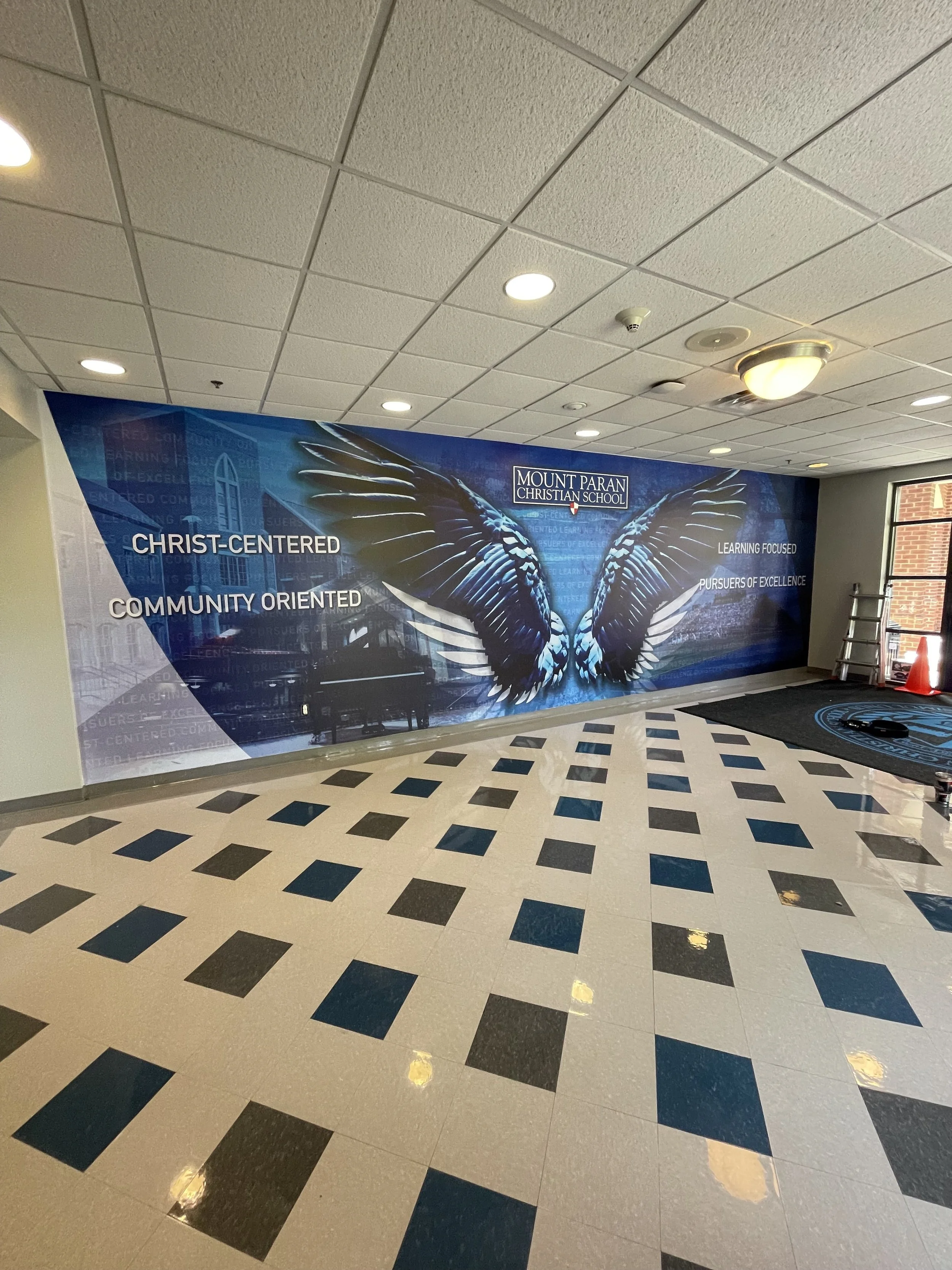 Interior of Mount Paren Christian School with a large blue banner featuring a butterfly graphic, school name, and motivational words on the wall, surrounded by tiled floor and ceiling lights.