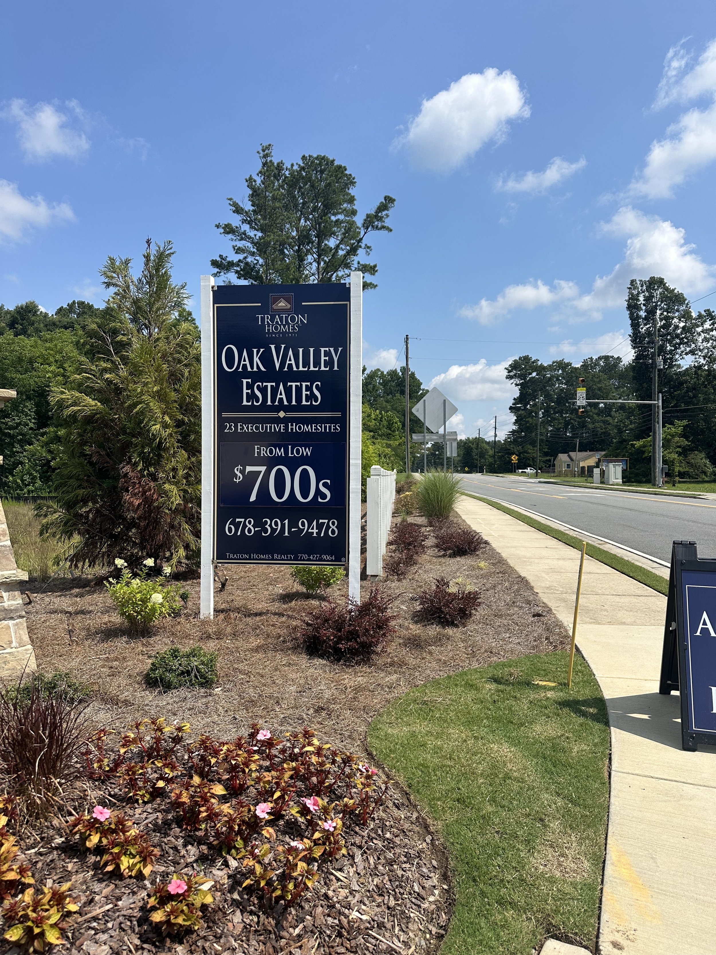 A street scene featuring a blue sign for Oak Valley Estates, a subdivision offering homes from $700s, with a decorated landscaped area with plants and flowers, a sidewalk, and a road with traffic signals and houses in the background, under a clear bl