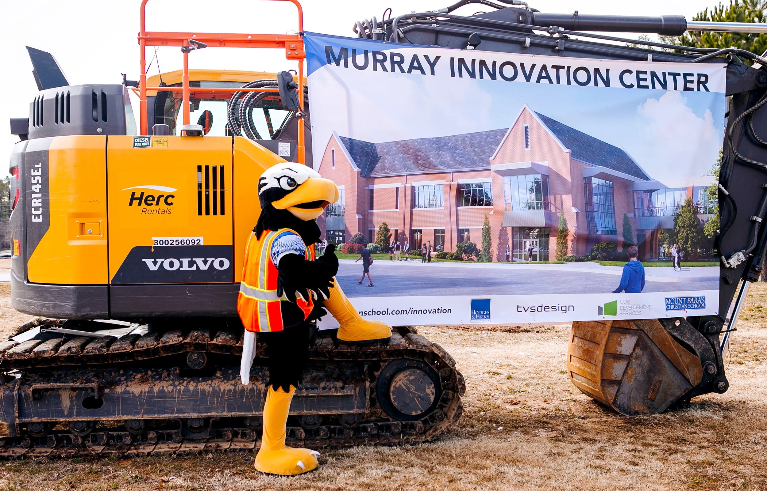 Construction site sign for Murray Innovation Center with an excavator in front, featuring a mascot dressed as a bird, in front of a building rendering.