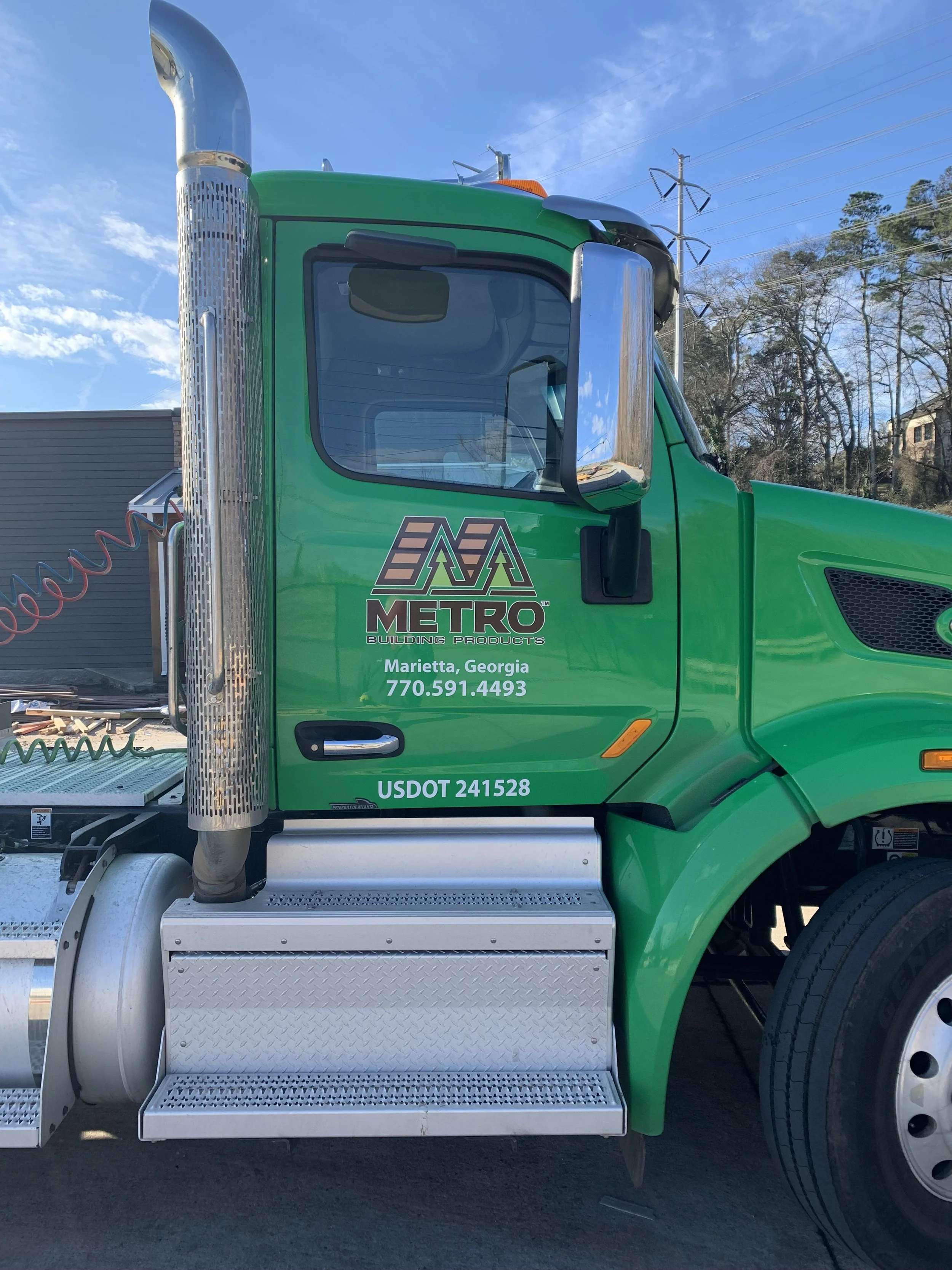 Green semi-truck with company logo and contact information on the door, chrome exhaust pipe, metal steps, black tires, and a background of trees, power lines, and a blue sky.