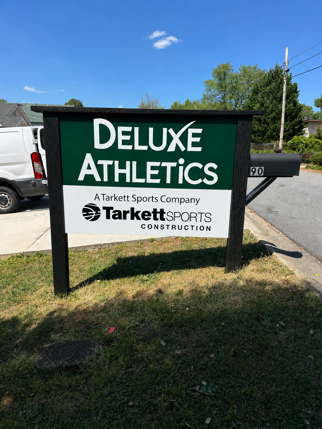 Sign for Deluxe Athletics, a Tarkett sports company specializing in sports construction, outdoors on a grassy area near a sidewalk, with a white van and trees in the background.