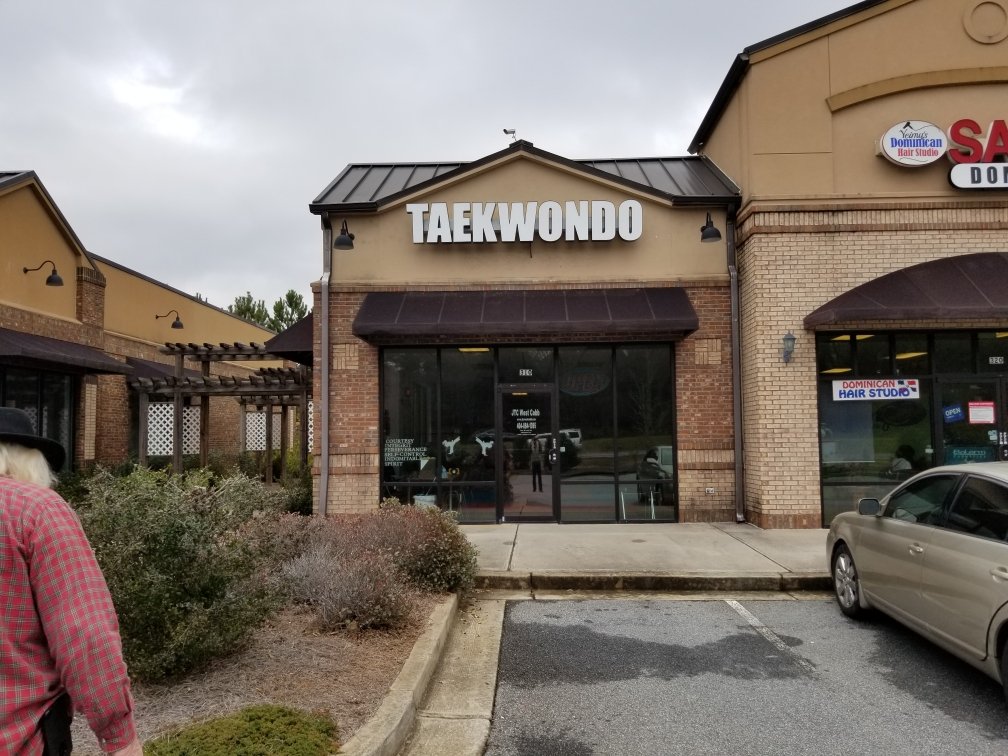Front of a retail building with a sign that reads 'TAEKWONDO', adjacent stores, a parking lot, and an overcast sky.