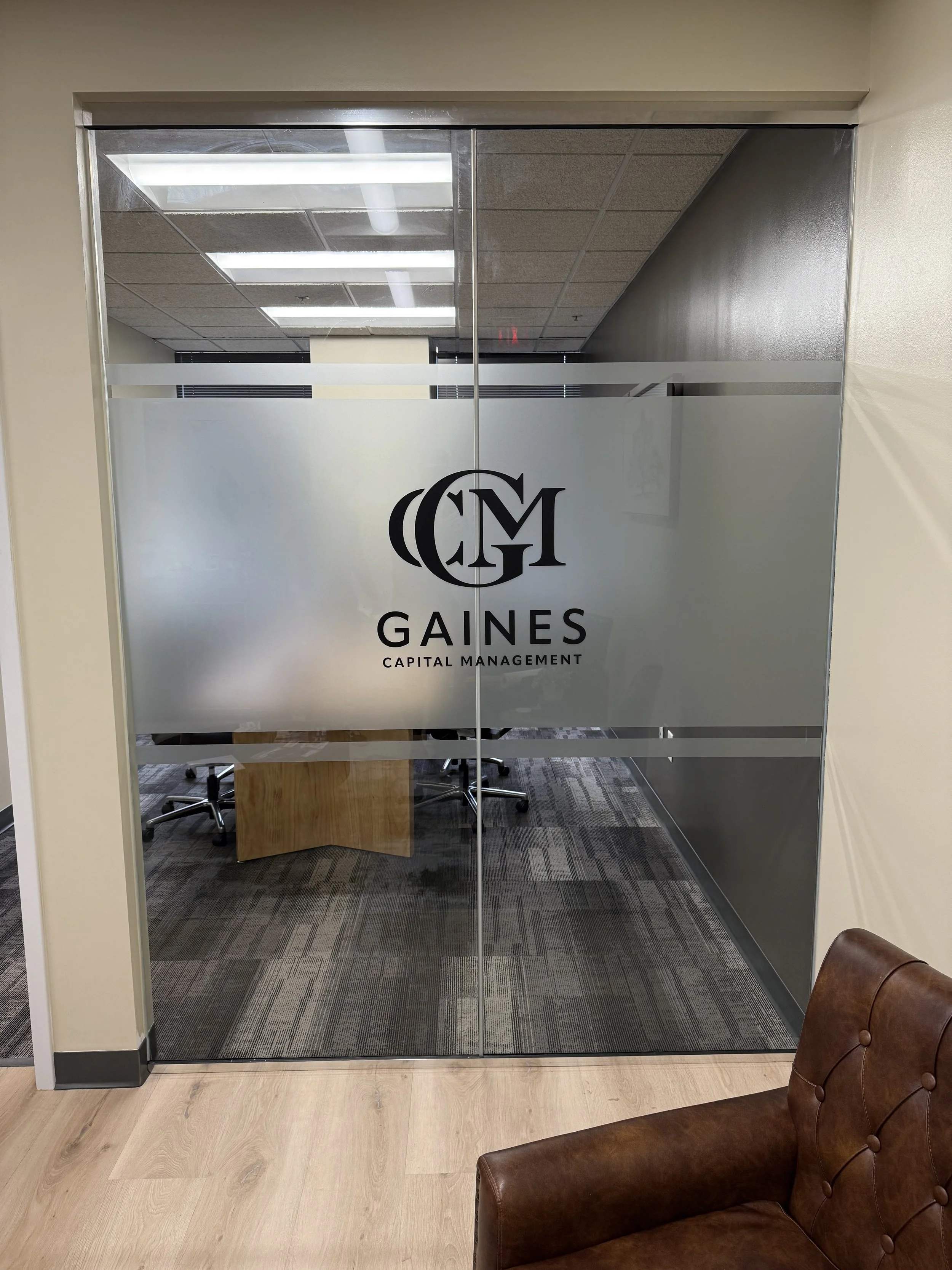 Office conference room with glass doors bearing the logo and name 'Gaines Capital Management' and a brown leather chair in the foreground.
