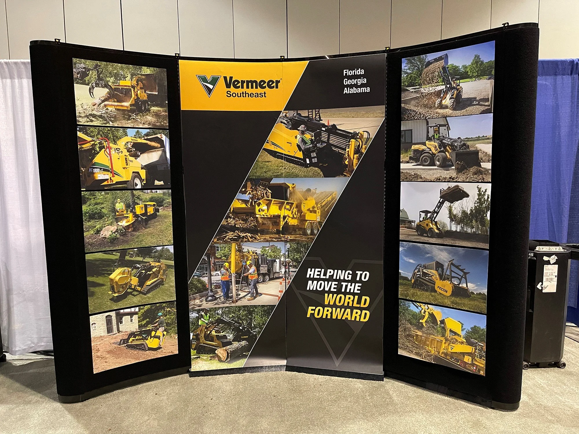 A display board showcasing various construction and landscaping equipment, with a logo for Vermeer Southeast and text indicating the regions Florida, Georgia, and Alabama, and the slogan "Helping to move the world forward." The board features multiple images of yellow machinery, including compact excavators, tree spades, and other heavy equipment, set against outdoor backgrounds.