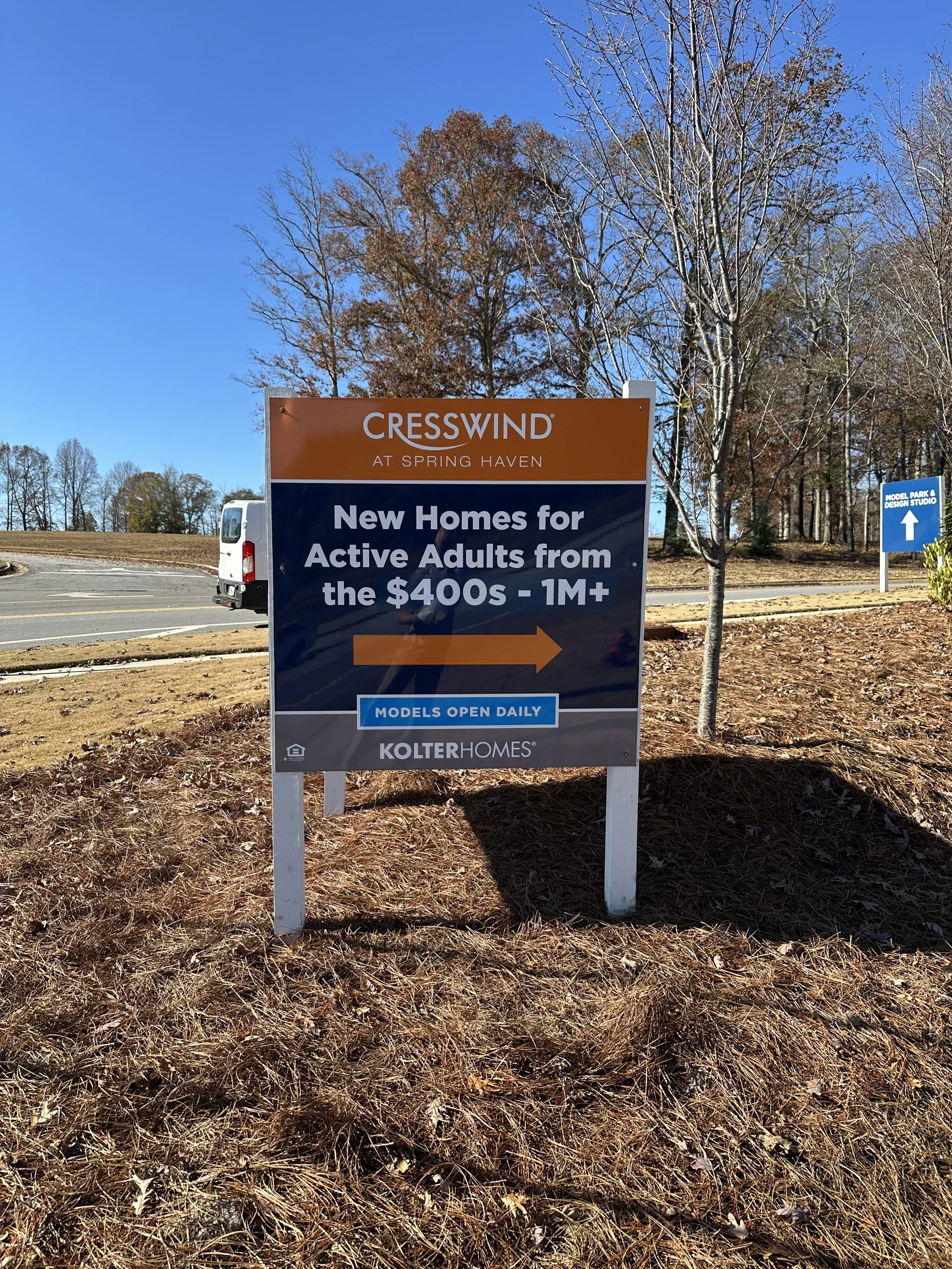 Sign at a new home development indicating model homes open daily, with a wooded area and a road in the background under a clear blue sky.