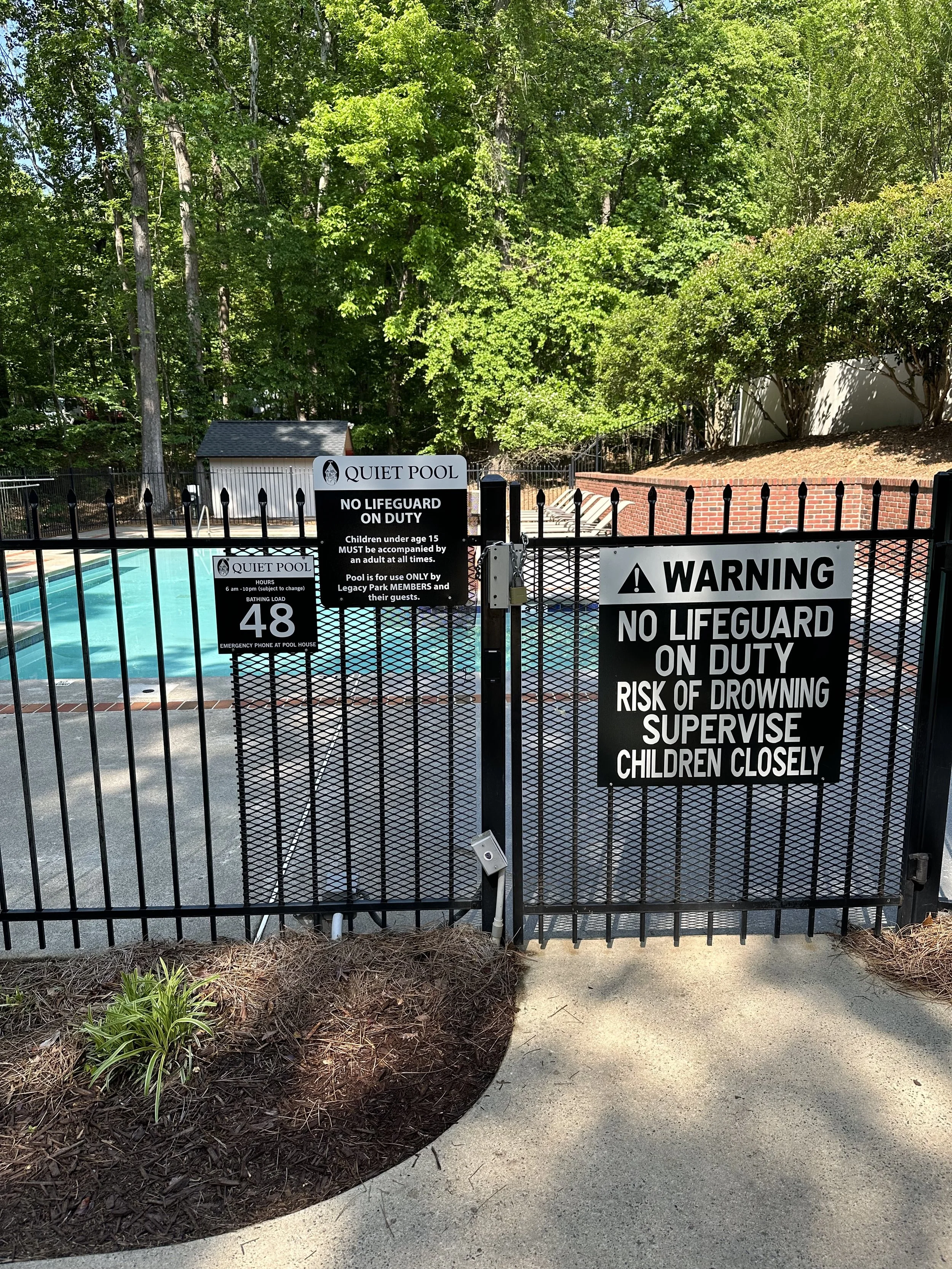 A fenced swimming pool area with signs. One sign says 'Quiet Pool, No Lifeguard on Duty, children under age 15 must be accompanied by an adult at all times, pool for use only by Legacy Park members and their guests.' Another large sign warns 'No Lifeguard on Duty, risk of drowning, supervise children closely.' The pool is outdoors, surrounded by trees and bushes, with lounge chairs and a small building in the background.