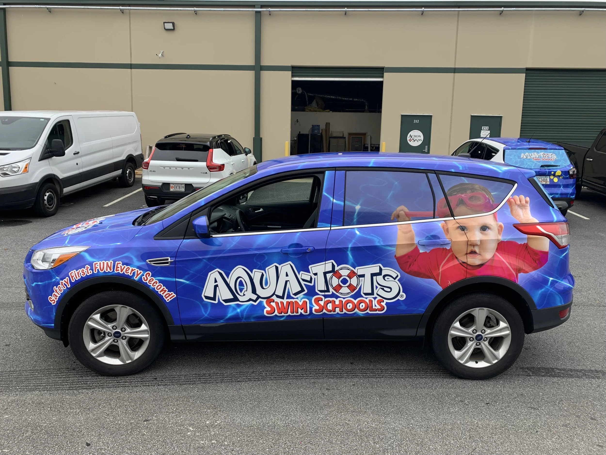 A blue car with a child holding a red swim noodle in the driver's seat, featuring a large graphic of a child in swimming goggles on the side. The background shows a parking lot with a white van, a white SUV, and a blue SUV in front of a beige industrial building. The blue SUV advertises Aqua-Tots Swim Schools.