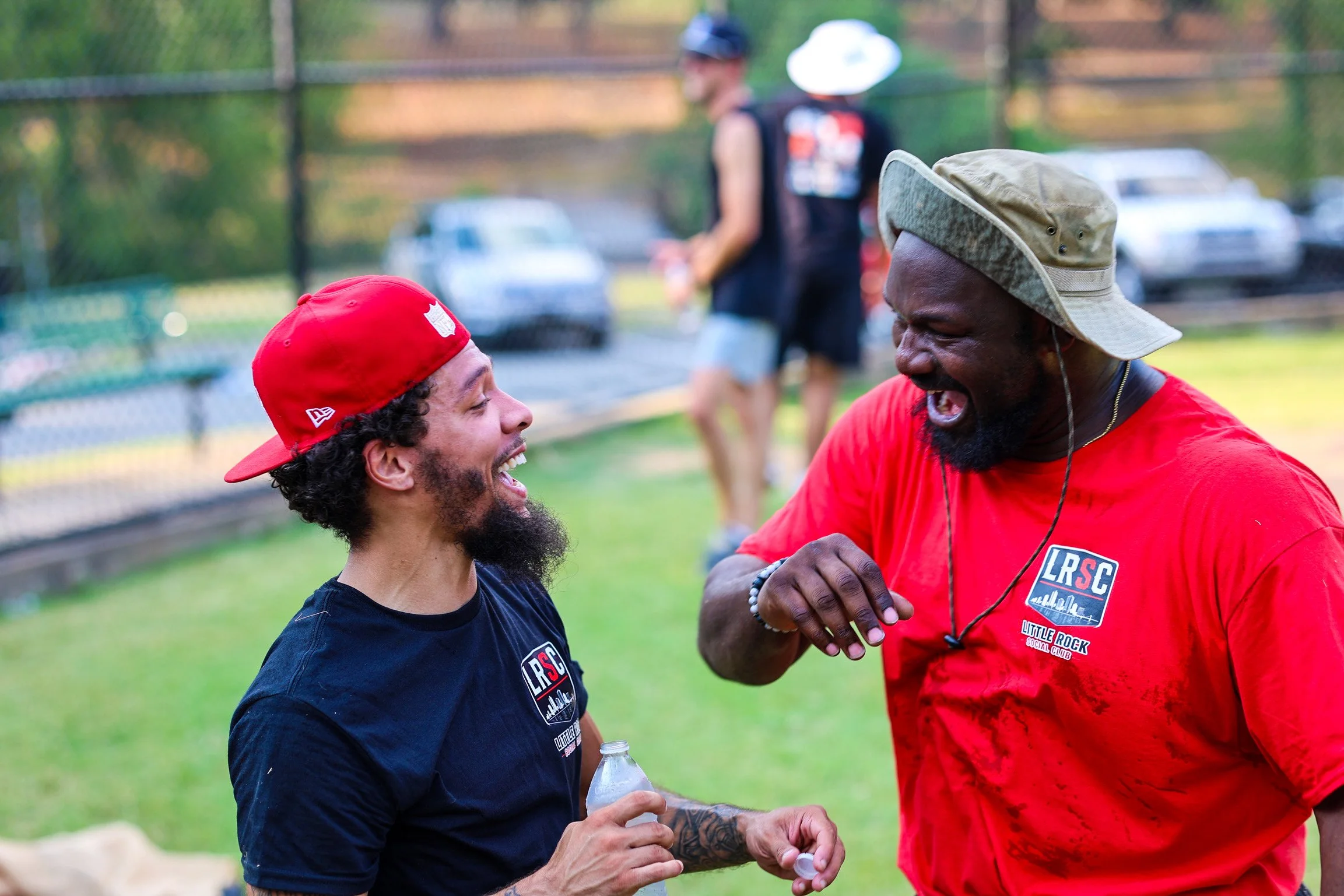 Two men laughing and talking outdoors near a park or sports field, with a person blurred in the background and several parked cars.