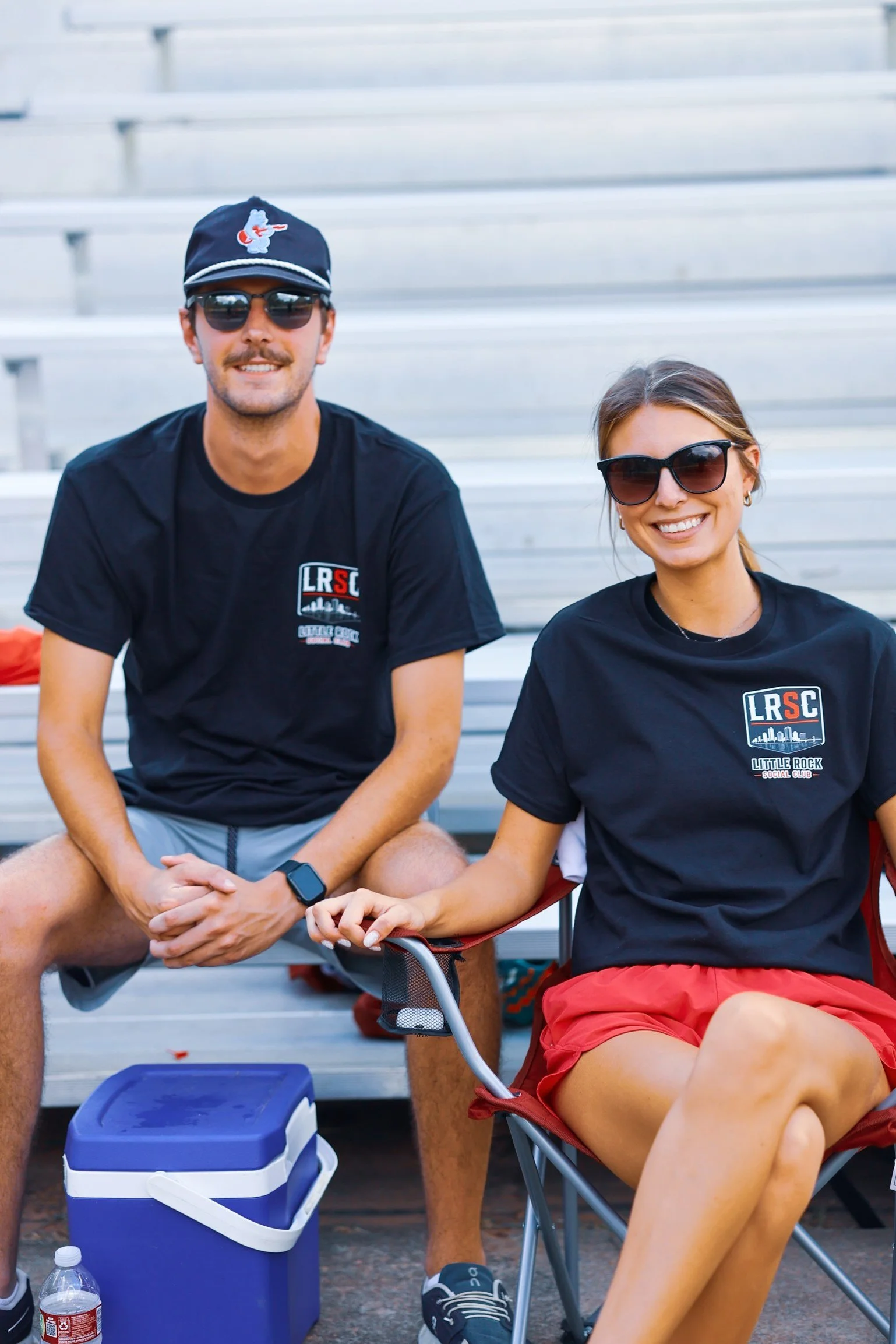 Two smiling people sitting on bleachers, wearing sunglasses and black t-shirts with a logo, outdoors.