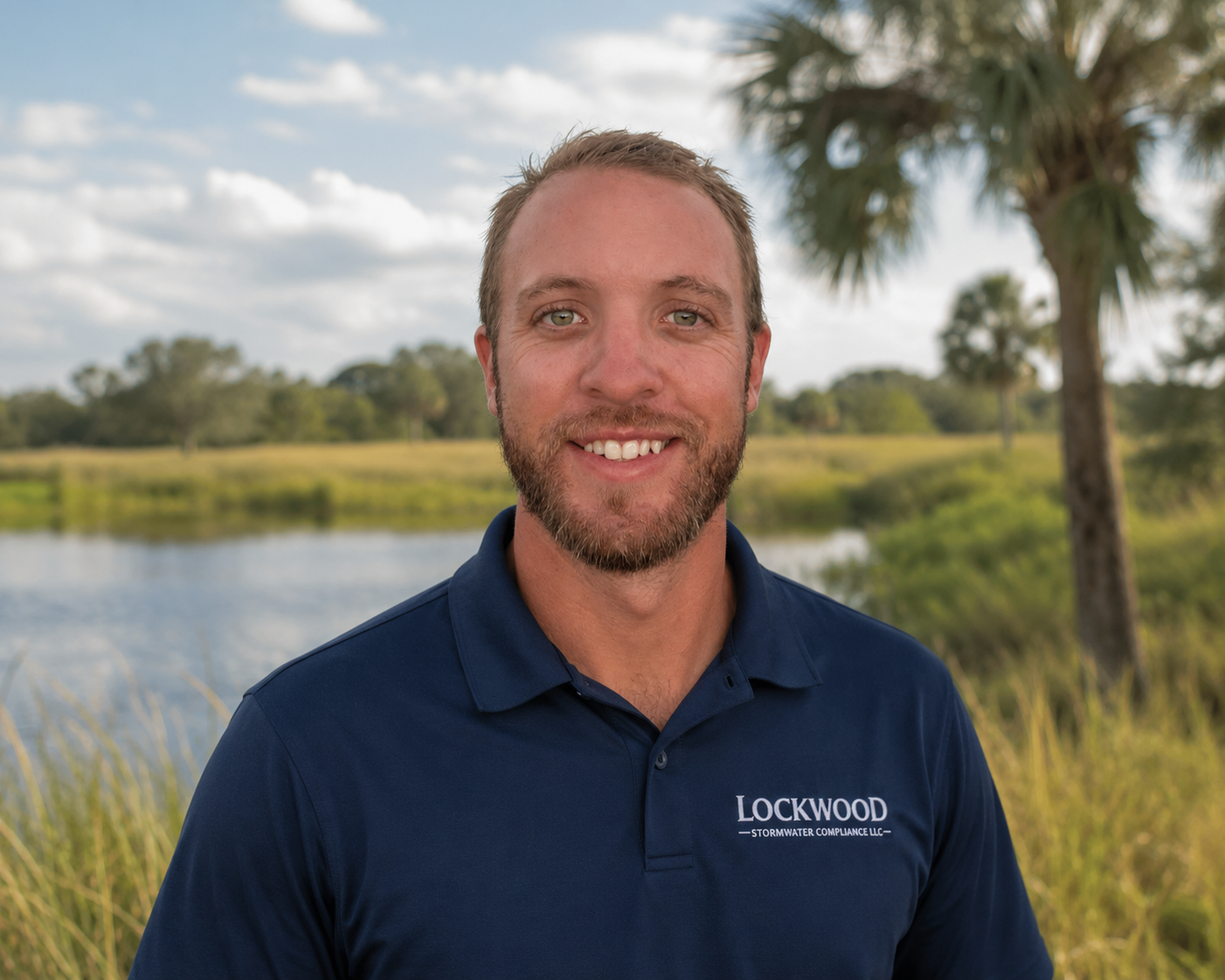 A smiling man in a navy polo shirt with 'Lockwood Stormwater Compliance LLC' embroidered, standing outdoors near water with trees, grass, and cloudy sky in the background.
