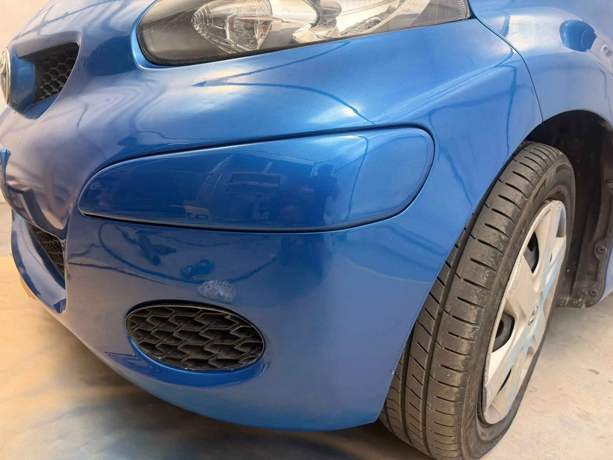 Close-up of the front left side of a blue sports car, showing the headlight, front grille, and part of the front wheel, all on a concrete floor.