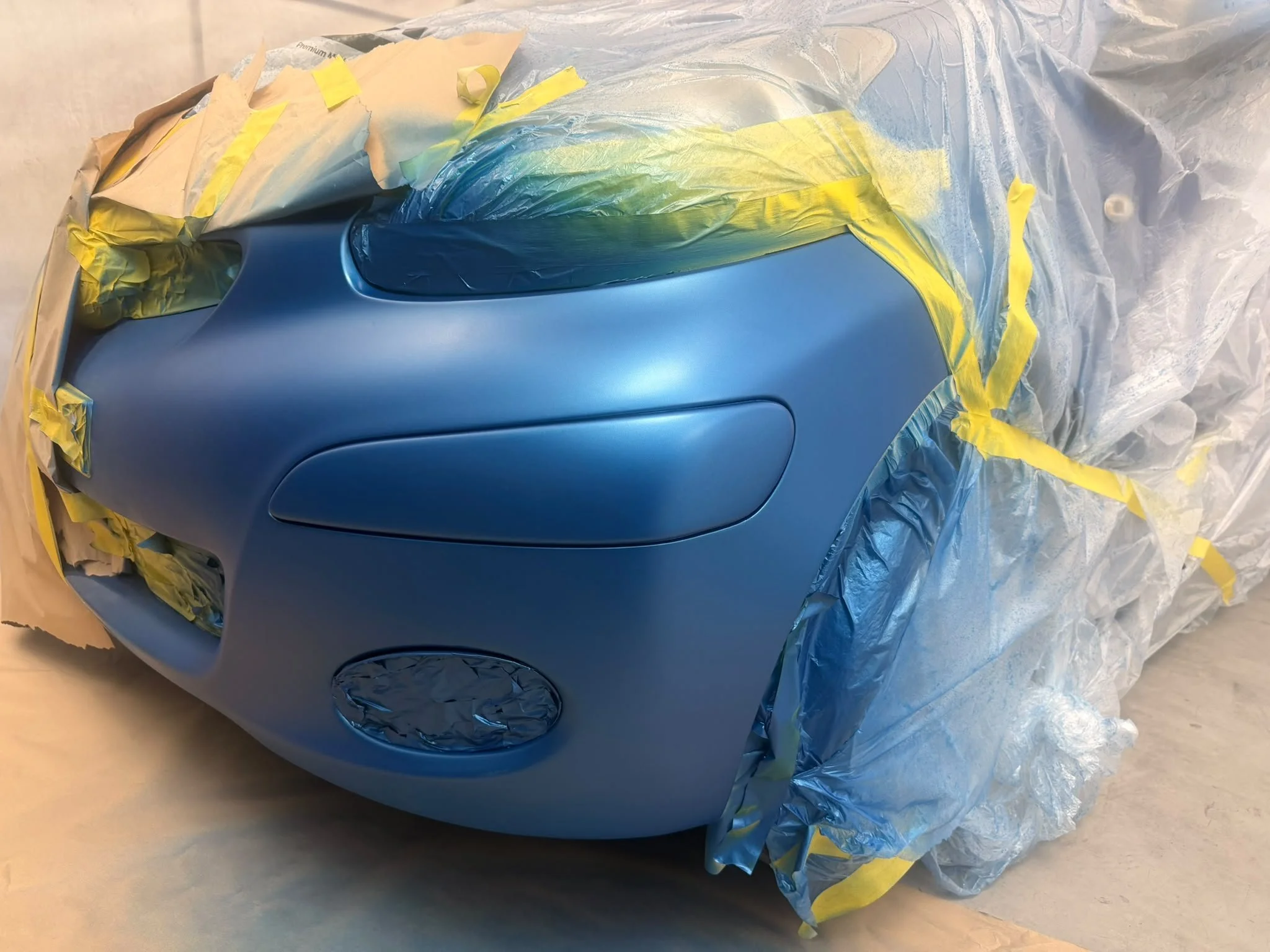 A car being painted blue surrounded by masking paper and yellow painter's tape.