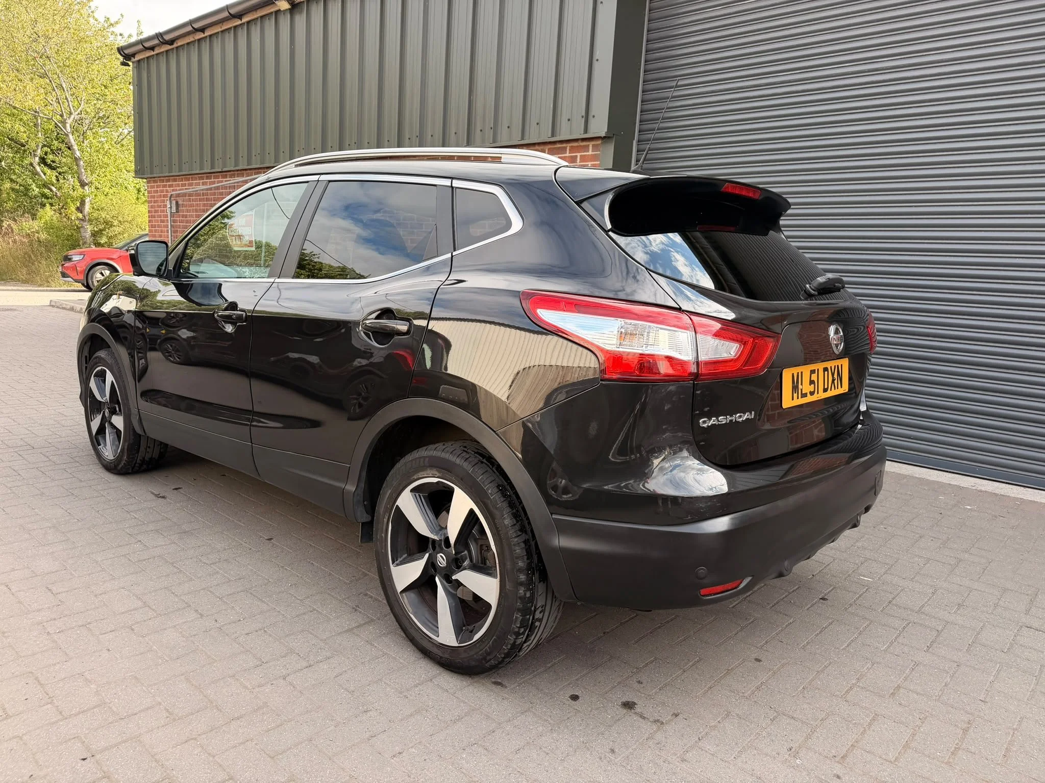 Black Nissan Qashqai parked in front of a building with a red brick wall and black and gray metal siding, with a red car partially visible in the background.